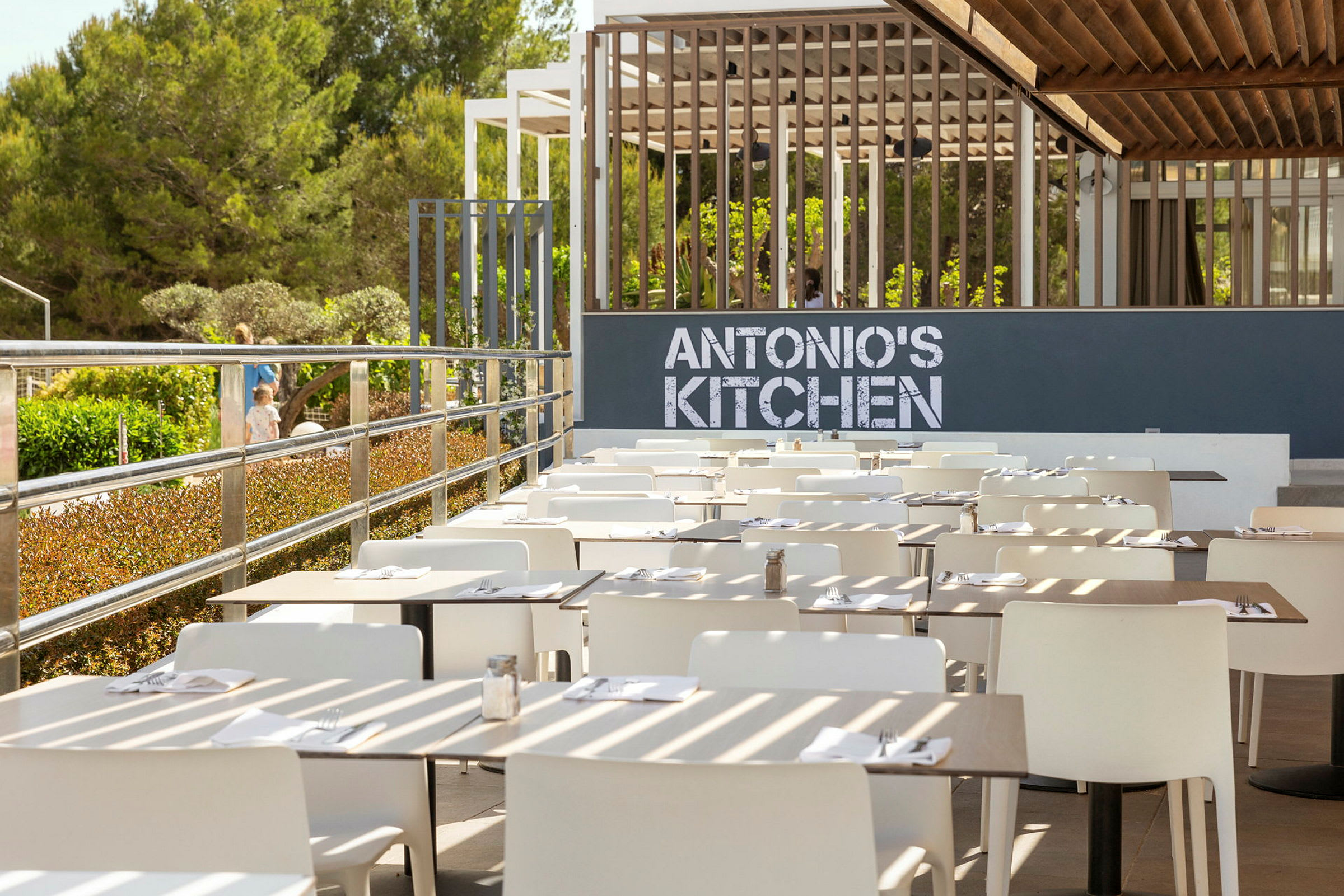 En uteservering med vita stolar och dukade bord framför en skylt med texten 'ANTONIO'S KITCHEN'. (AI-genererad text)