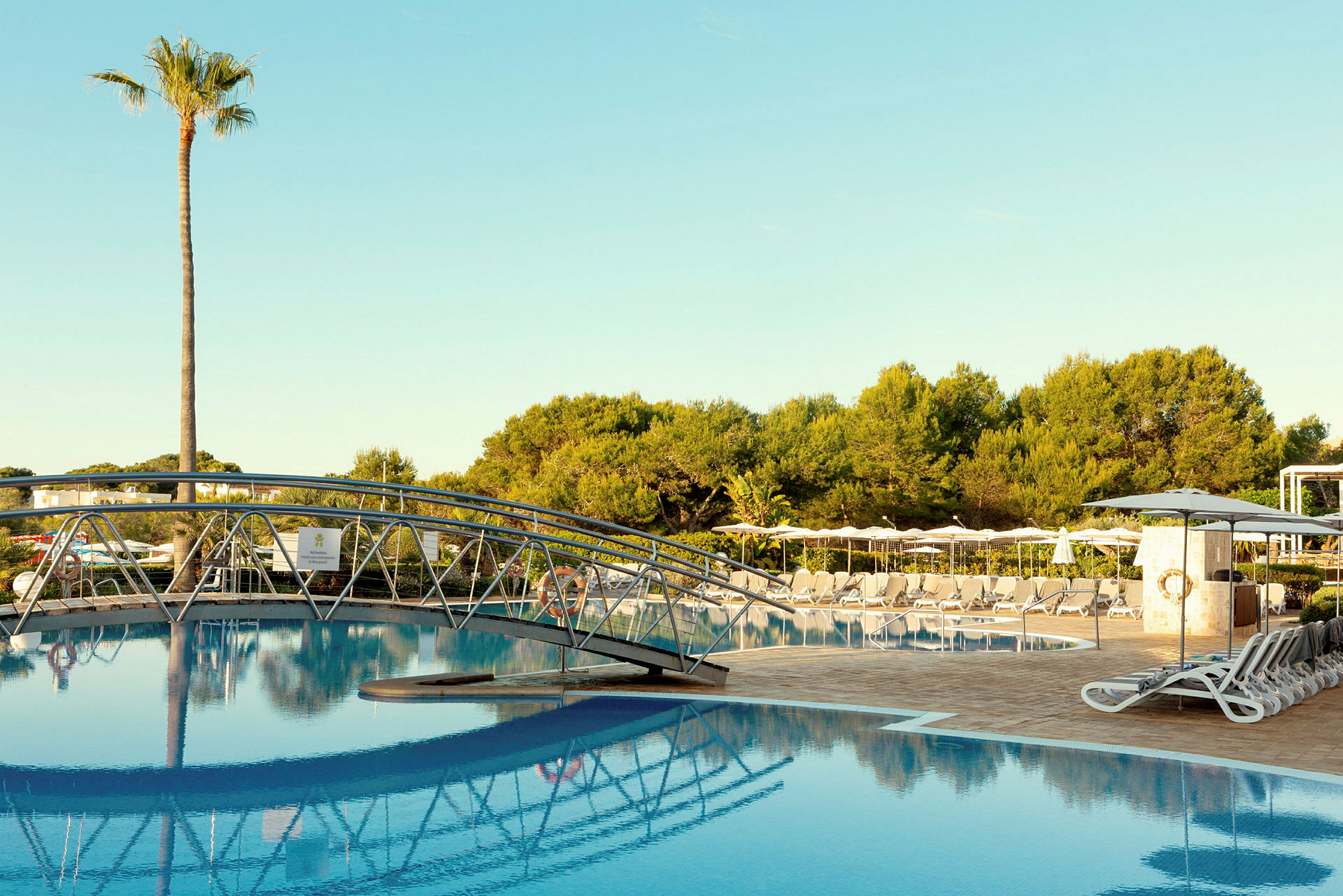 Pool med en bro i förgrunden, solstolar och parasoller runt om samt träd i bakgrunden. (AI-genererad text)