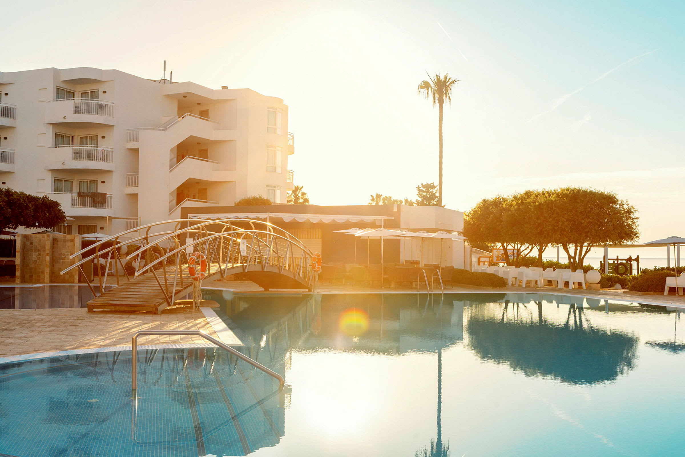 Bild av en utomhuspool vid ett hotell med en bro över poolen, omgivna av grönska och byggnader. (AI-genererad text)