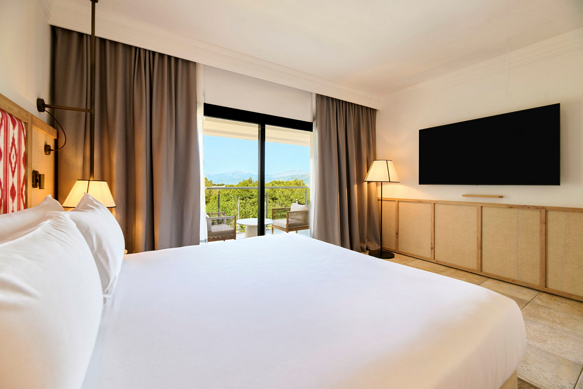 Ett hotellrum med en stor säng, TV på väggen, och en balkong med utsikt över grönt landskap. (AI-genererad text)