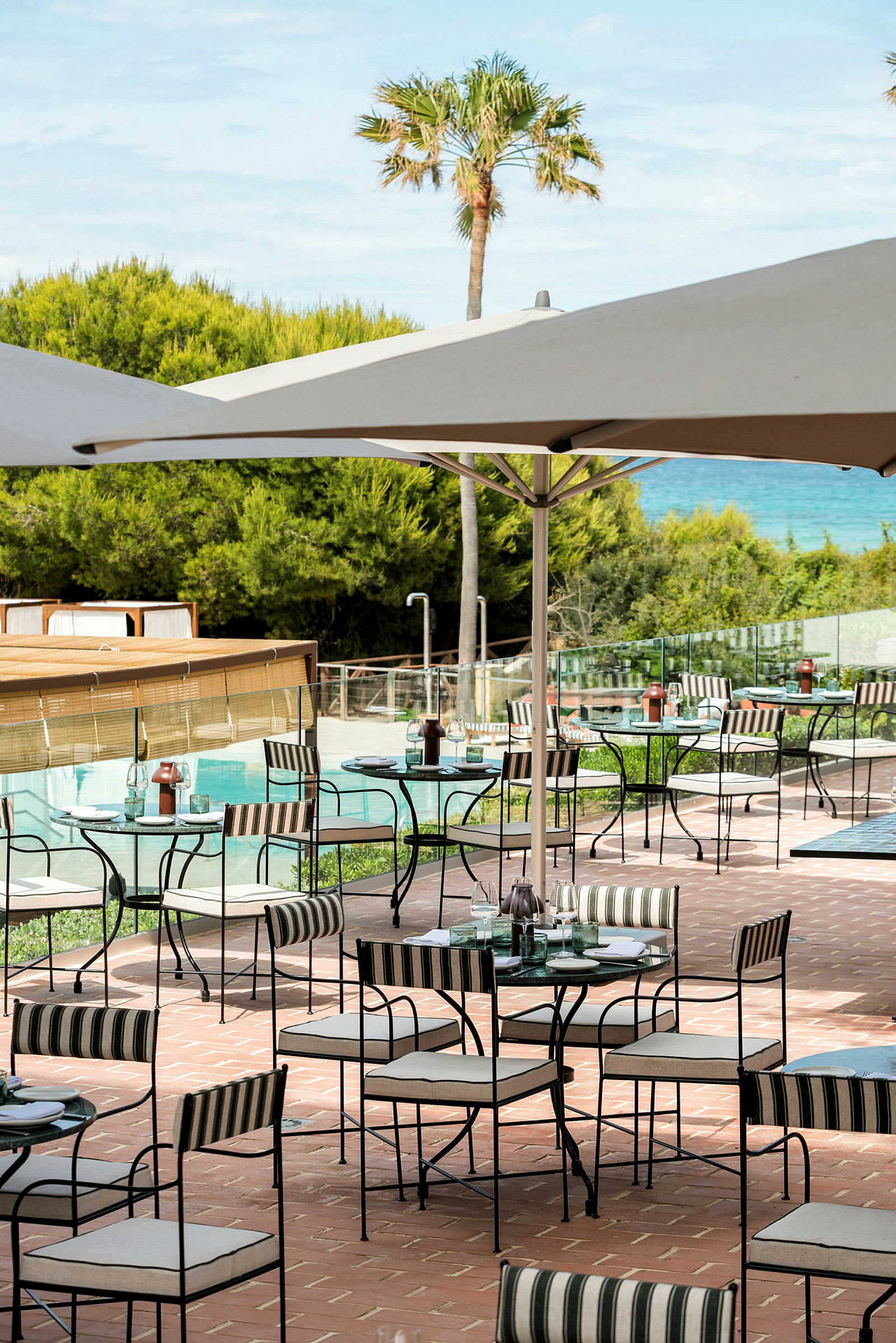 En uteservering med bord och stolar under parasoller på en terrass med utsikt över grönska och en pool. (AI-genererad text)