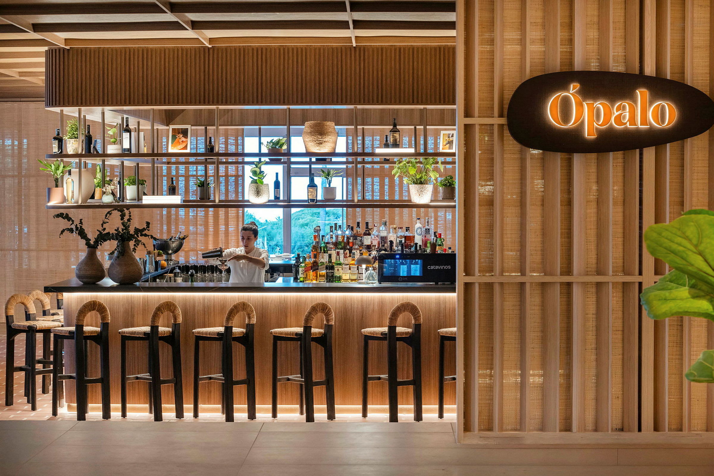 En elegant bar med trädetaljer och växter, där en bartender förbereder en dryck. (AI-genererad text)