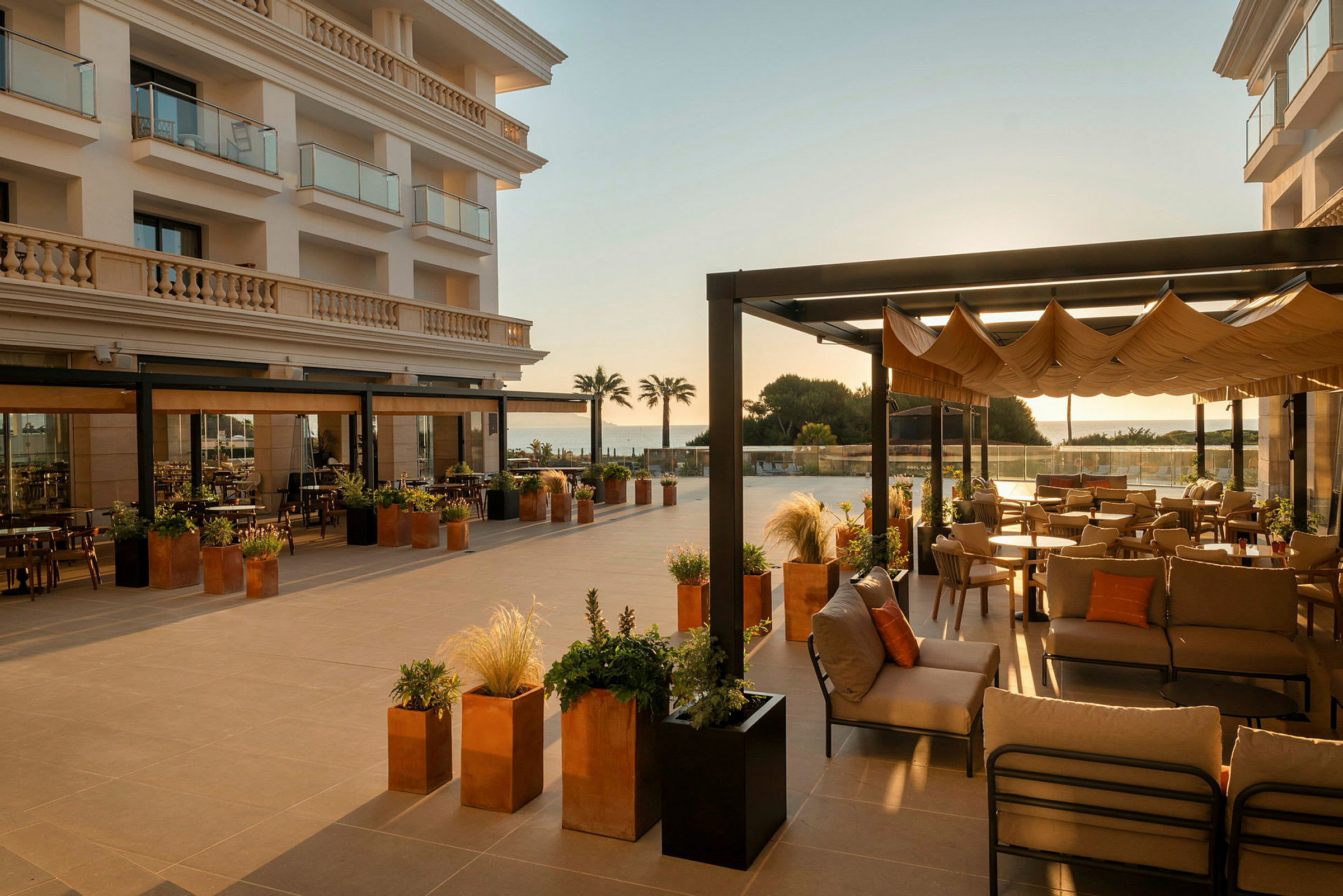 En rymlig uteservering vid ett hotell är inredd med komfortabla möbler och har utsikt över havet. Detaljer som växter i krukor och pergolor stärker den inbjudande stämningen. (AI-genererad text)