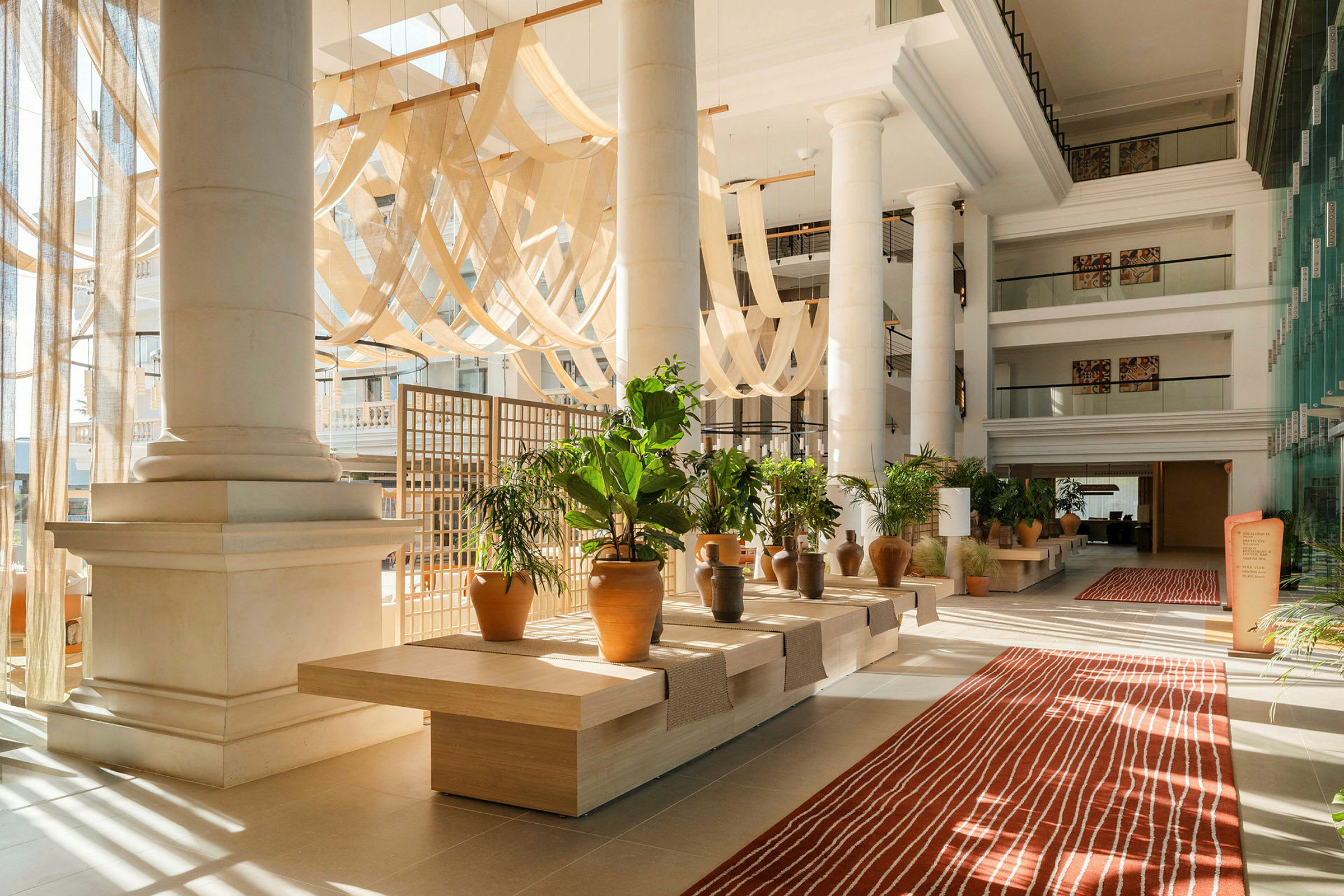 Ljus och luftig hotellobby med stora pelare, dekorativa växter och en röd matta. (AI-genererad text)
