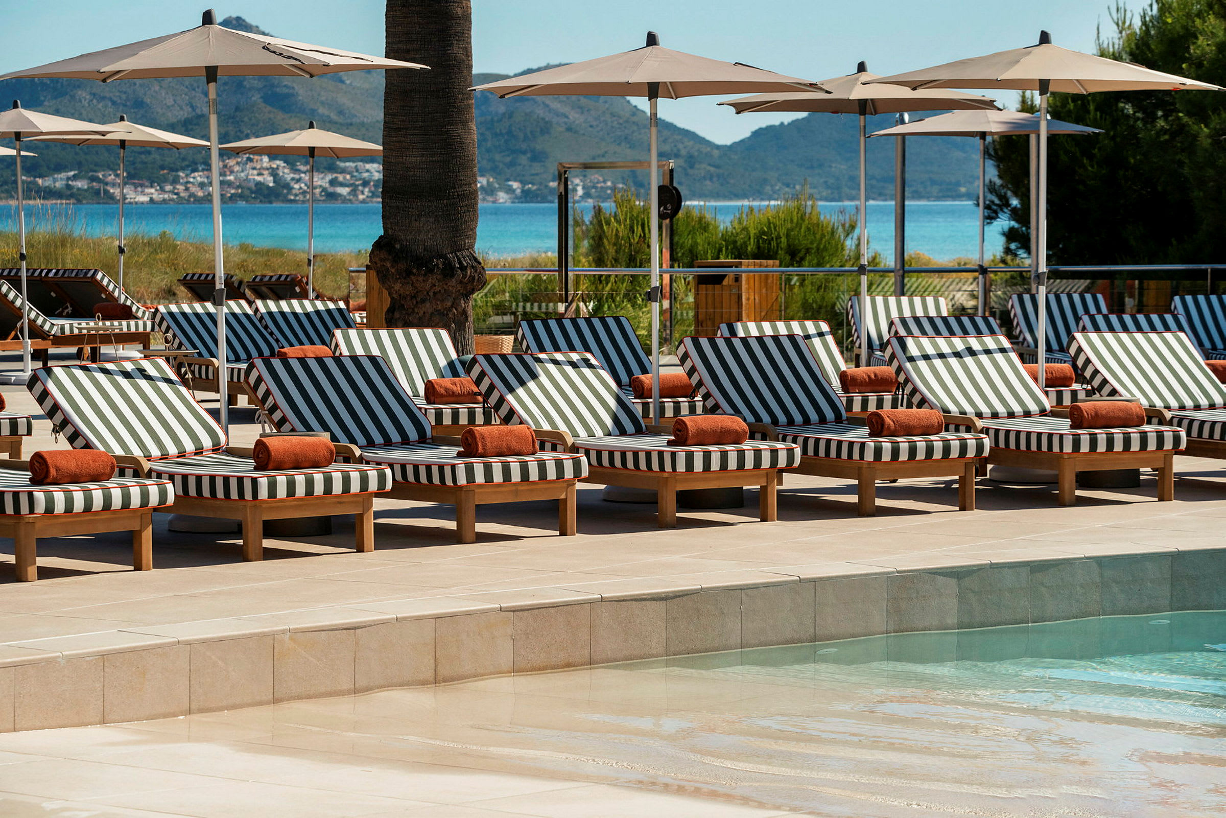 Solstolar med randiga dynor och parasoll vid en pool, med utsikt över havet och träd i bakgrunden. (AI-genererad text)