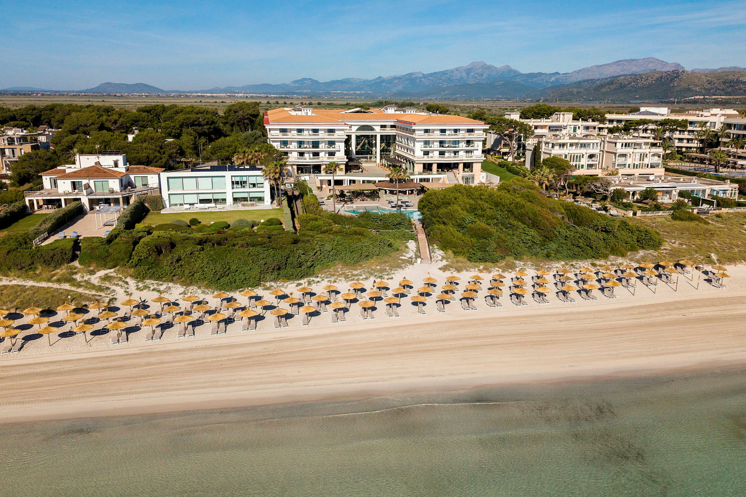 Flygfoto av ett hotellkomplex med en sandstrand och parasoller i förgrunden. (AI-genererad text)
