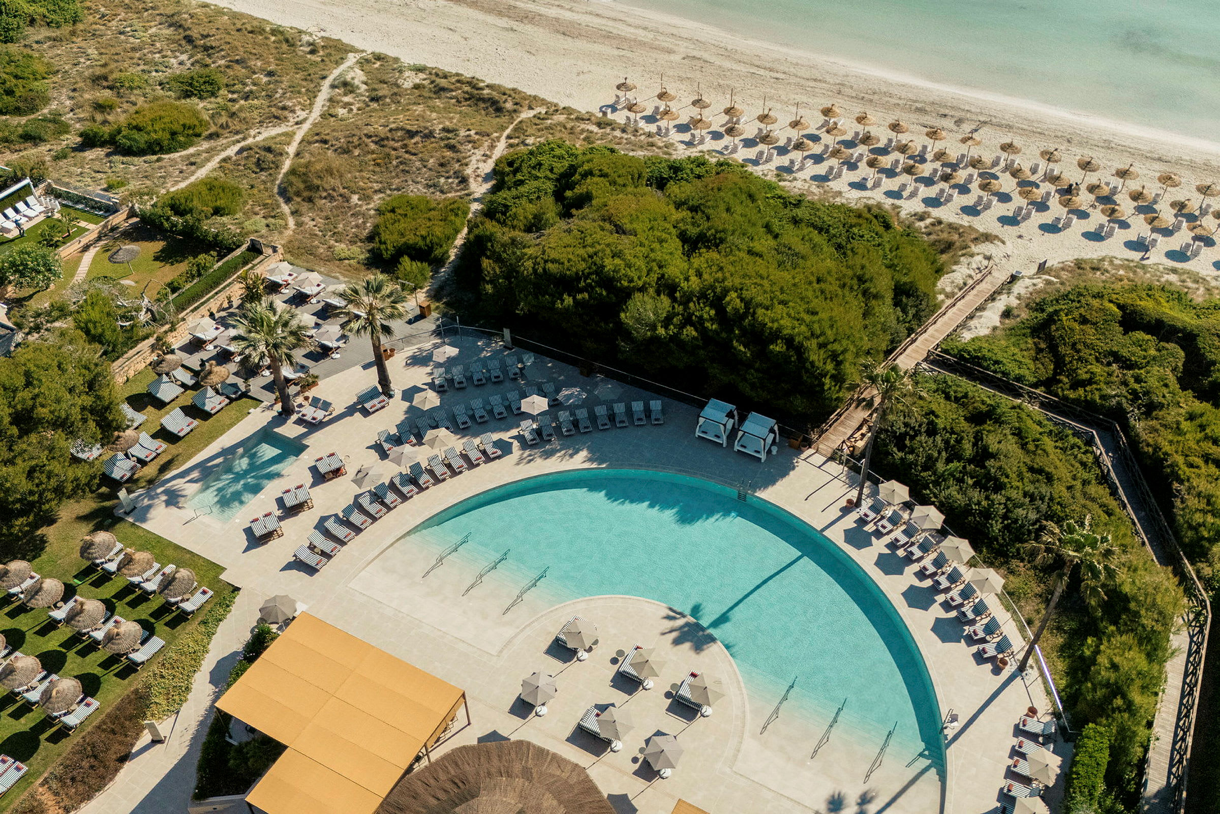 Flygvy över en pool, solstolar och parasoll, omgiven av grönska och en sandstrand i bakgrunden. (AI-genererad text)