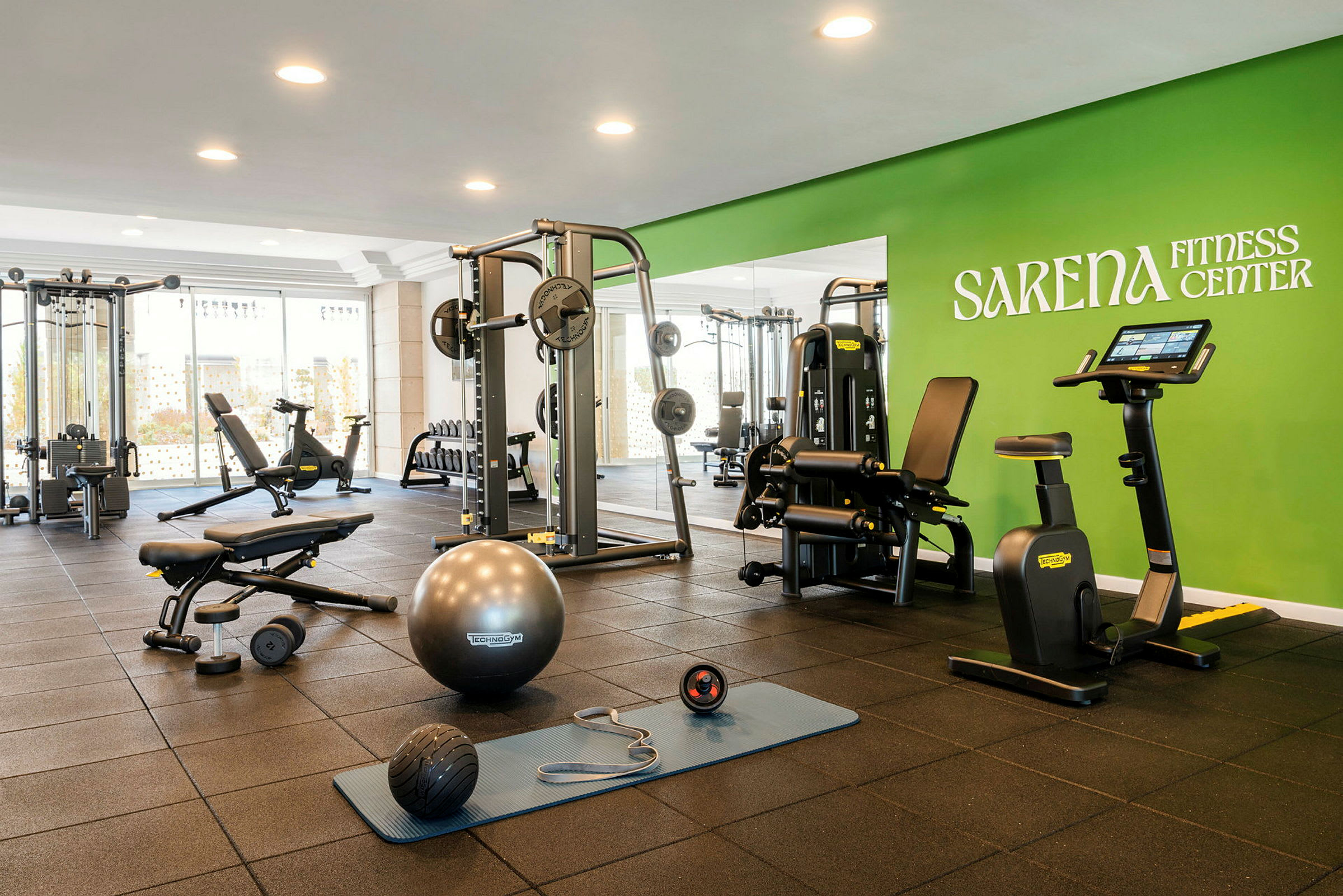 Ett gym med olika träningsmaskiner och redskap, inklusive motionscykel, hantlar och en träningsmatta, samt grön vägg med texten Sarena Fitness Center. (AI-genererad text)