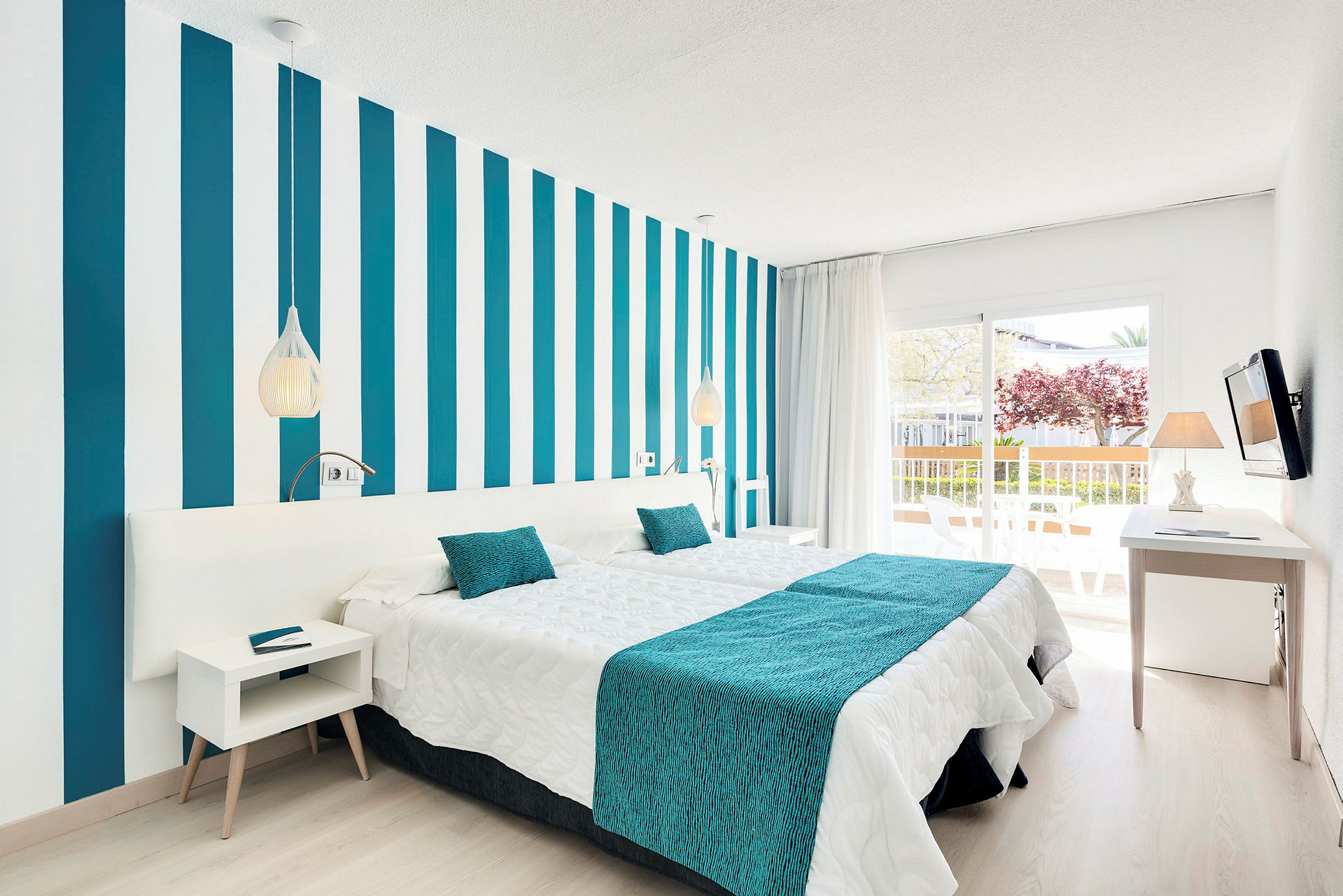 Ett modernt hotellrum med randig vägg, vita och turkosa sängkläder samt en balkong med utsikt. (AI-genererad text)
