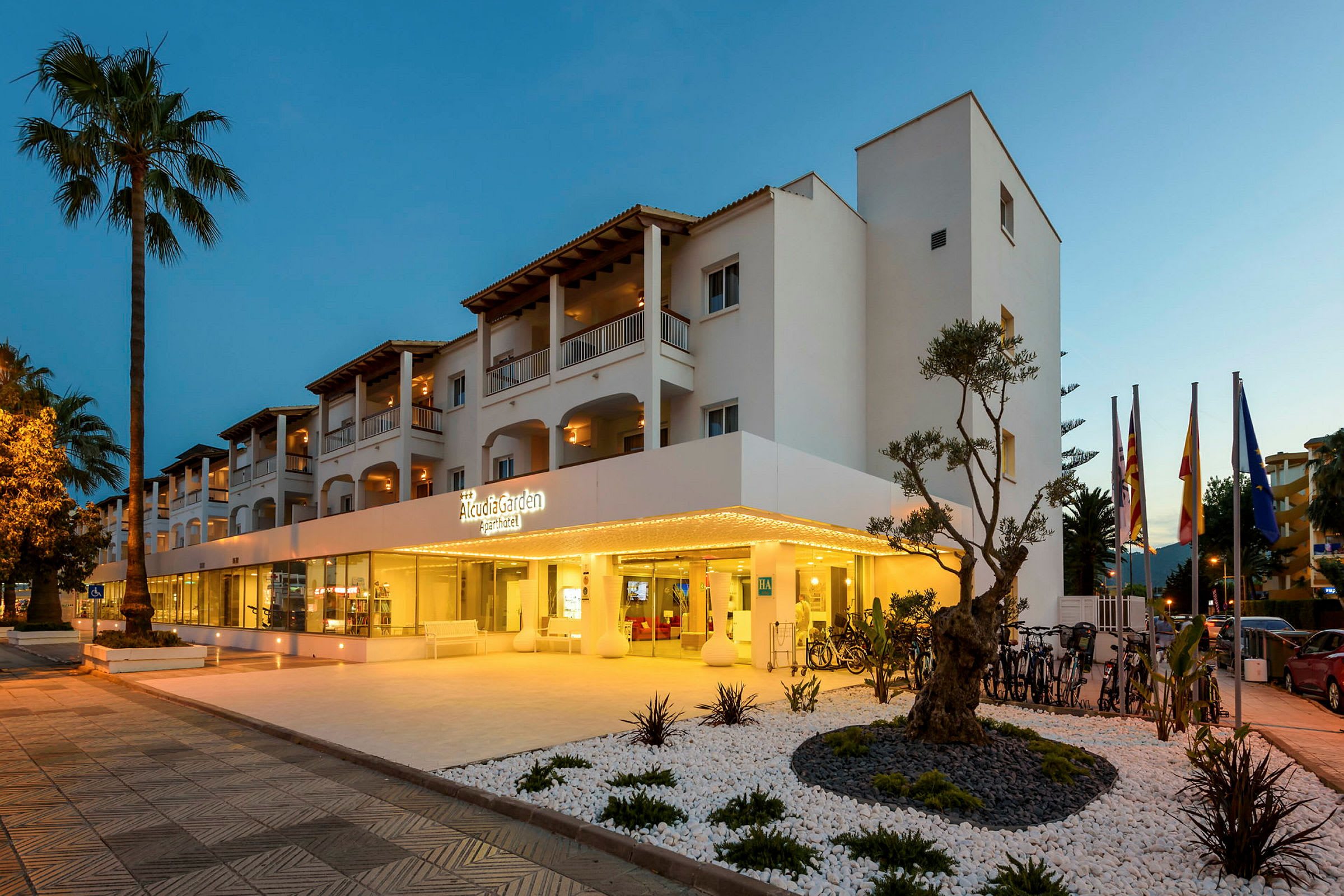 Bild av hotellet Alcudia Garden Aparthotel, med upplyst entré och omkringliggande trädgård. (AI-genererad text)