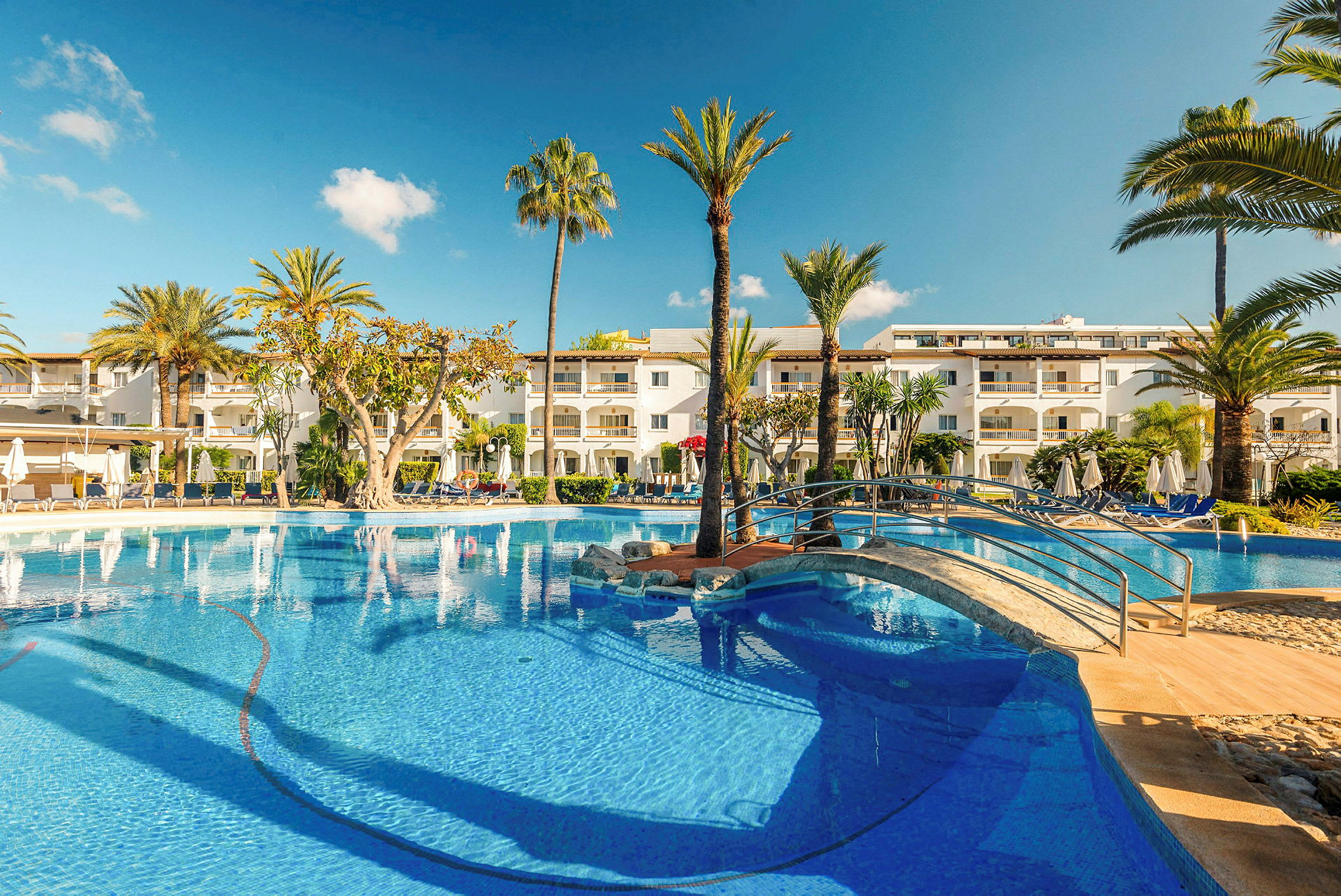 Utsikt över ett hotellområde med pool, omgiven av palmer, solstolar och grönska. (AI-genererad text)