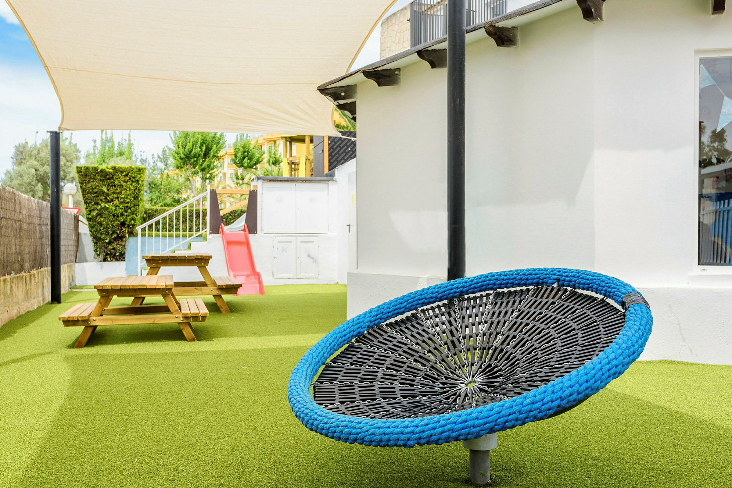 En lekplats med grön konstgräs, sittplatser vid träbänkar, en röd rutschkana och en blå hänggunga. (AI-genererad text)