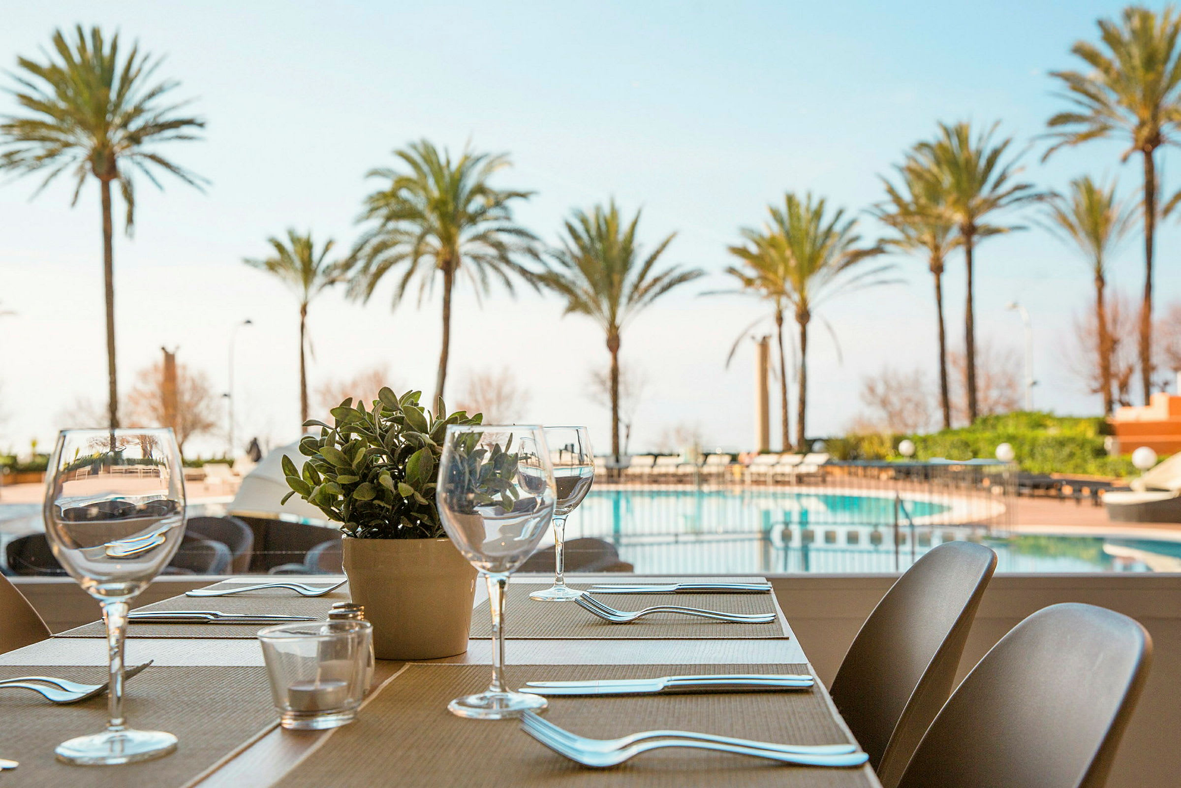 En dukad restaurangbord vid fönstret, med utsikt över en pool och palmer utomhus. (AI-genererad text)