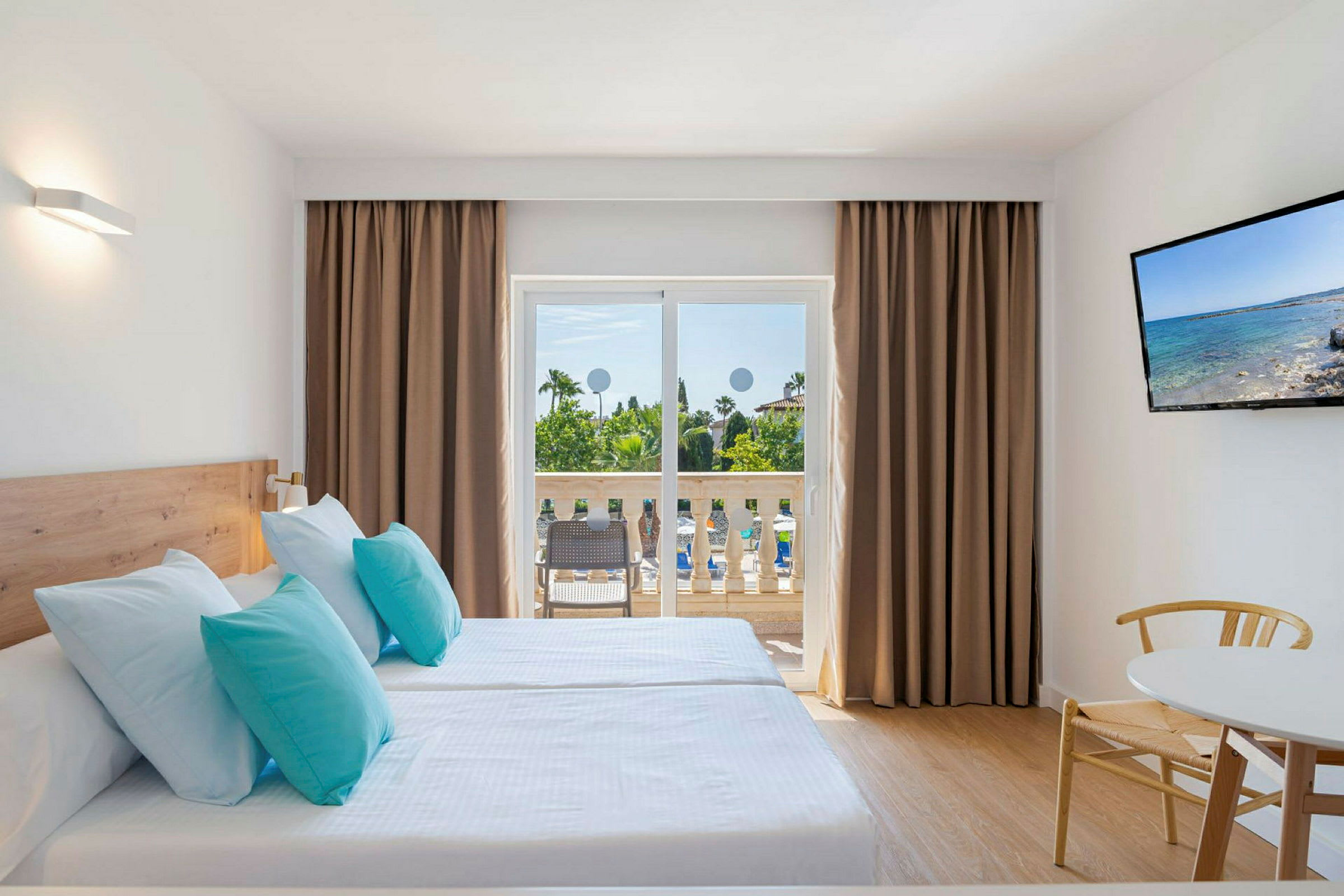 Ett hotellrum med två enkelsängar med ljusblå och turkosa kuddar, en balkong med utsikt över trädgård, och en TV på väggen. (AI-genererad text)