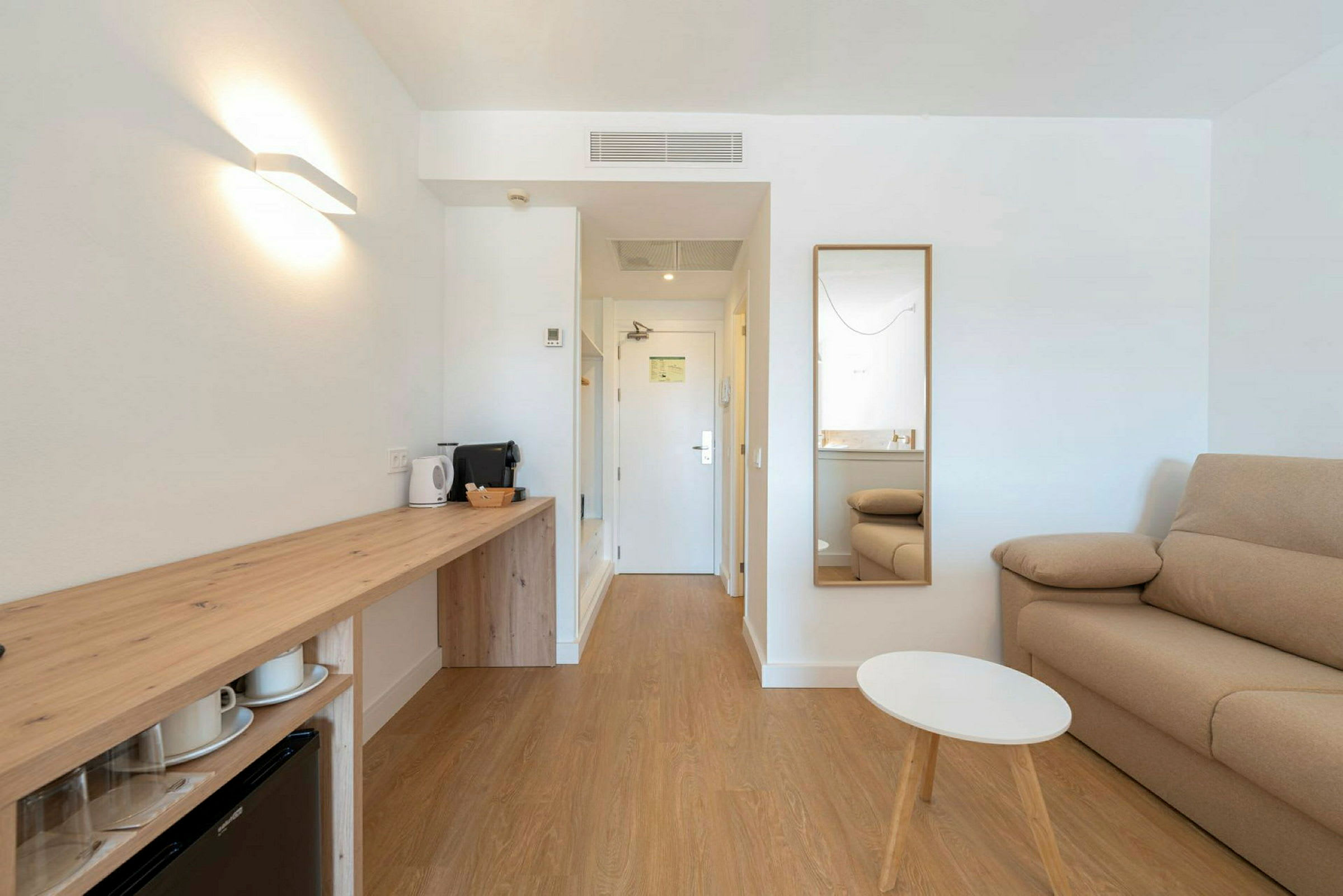Ett modernt hotellrum med en soffa, spegel, litet bord och en arbetsyta med vattenkokare och kaffemaskin. (AI-genererad text)