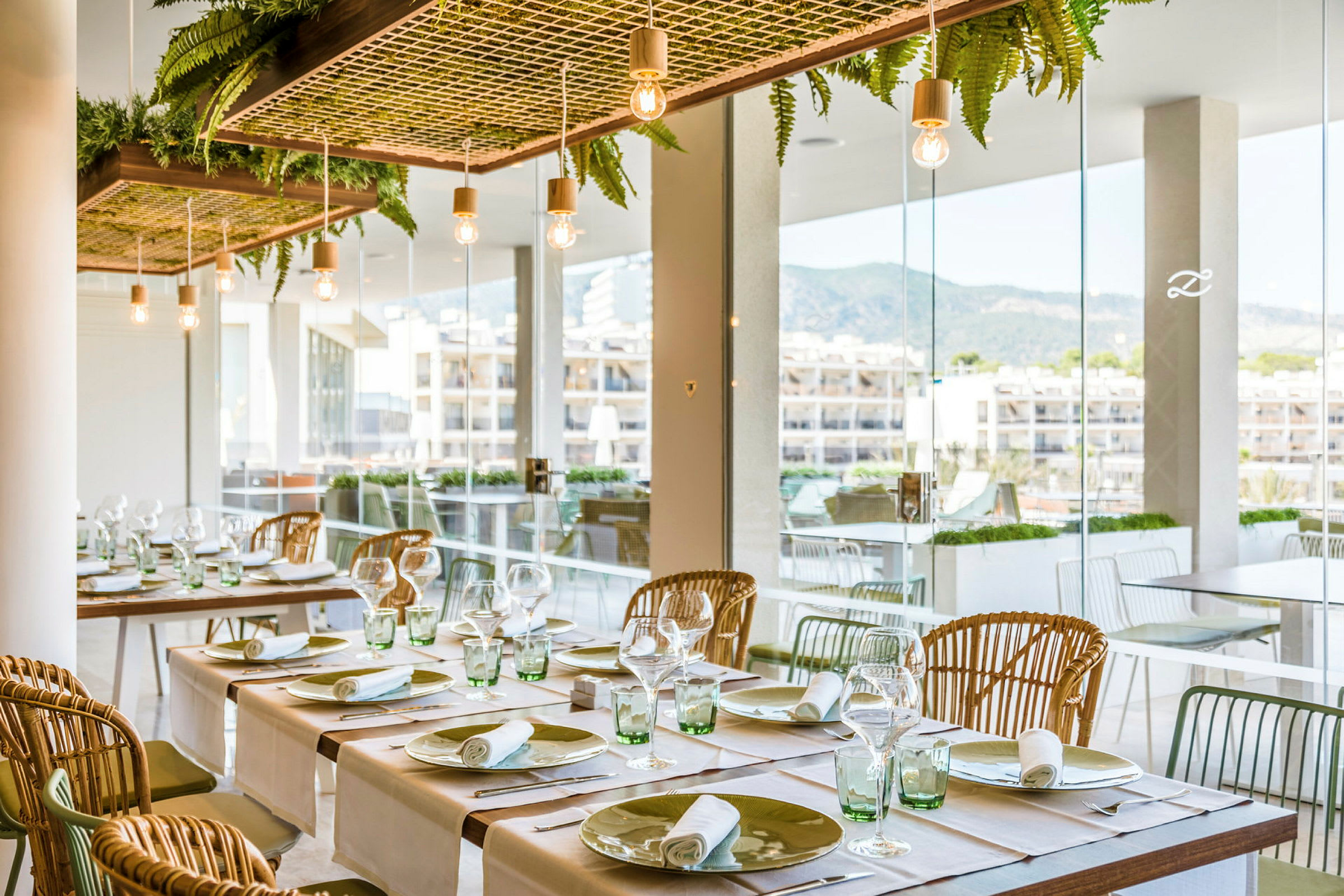 Interiör av ett restaurangområde med dukade bord, en modern inredningsdetalj med hängande växtdekorationer och glasväggar som ger utsikt över byggnader och berg. (AI-genererad text)