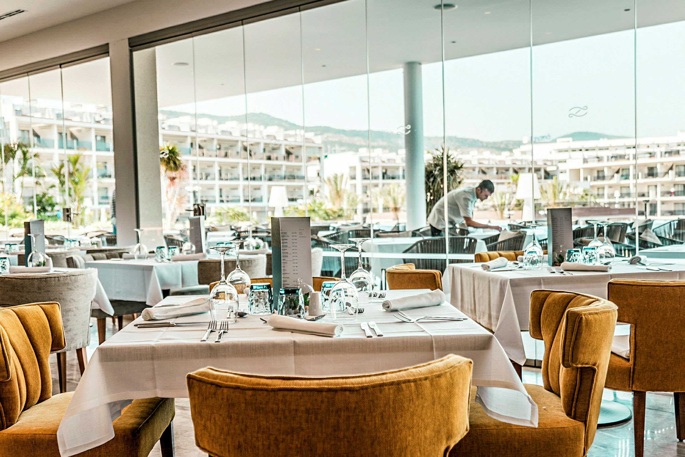 En modern restaurang med dukade bord och stolar samt fönster med utsikt över ett hotellområde. (AI-genererad text)