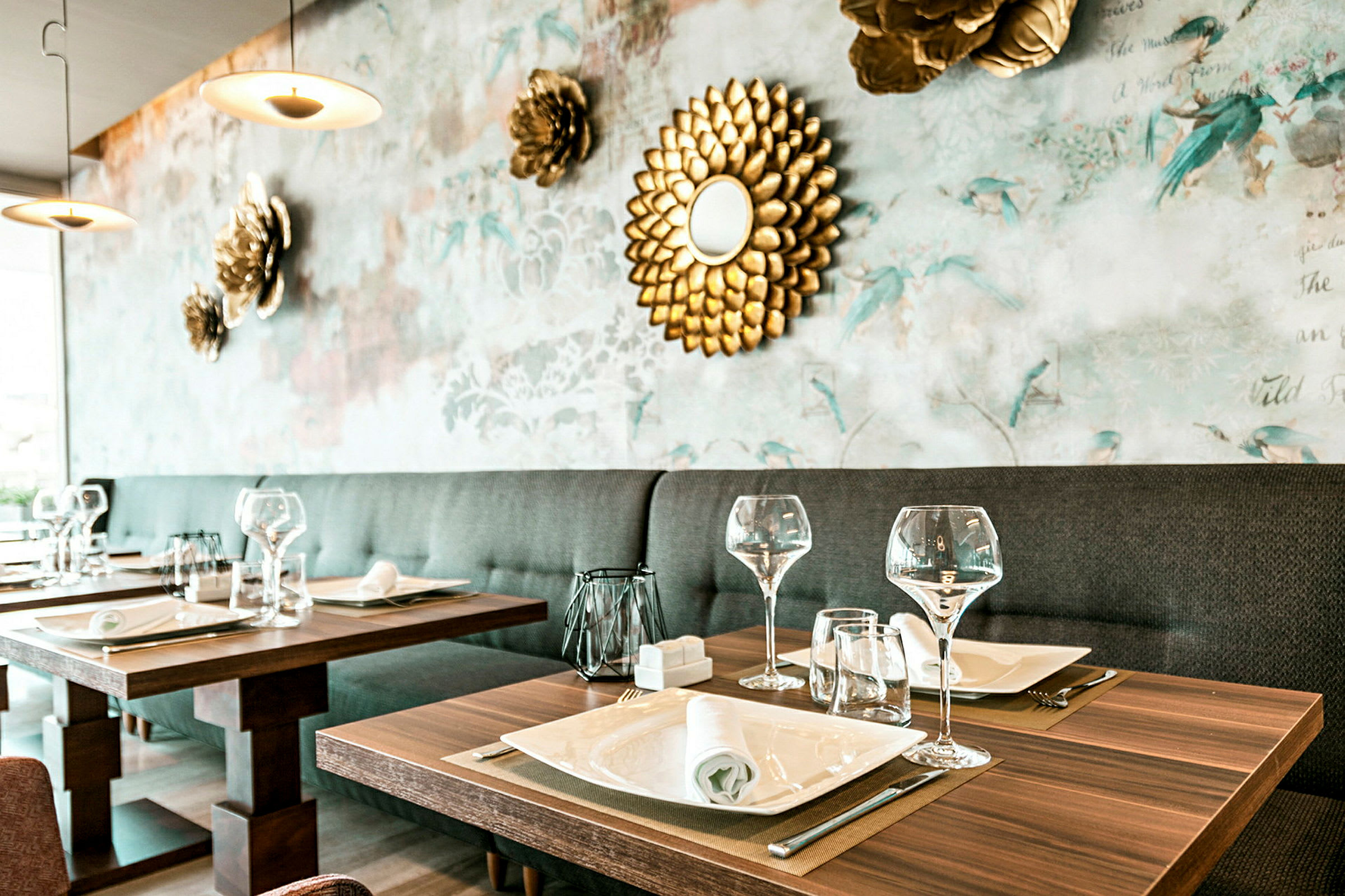 Ett elegant restaurangbord dukat med glas, tallrikar och servetter, med en dekorativ bakgrundsvägg prydd med blommor och en gyllene spegel. (AI-genererad text)