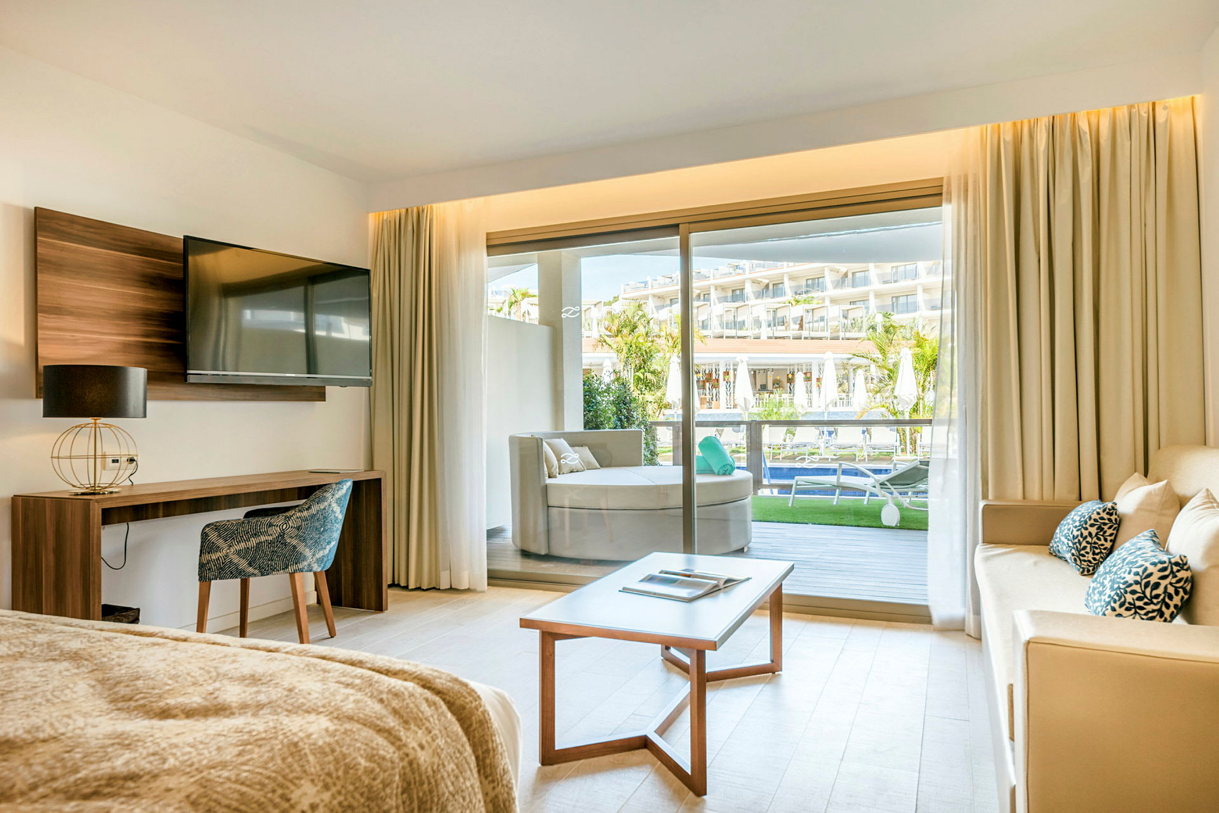 Ett hotellrum med soffa, bord och TV samt utsikt över en balkong. (AI-genererad text)
