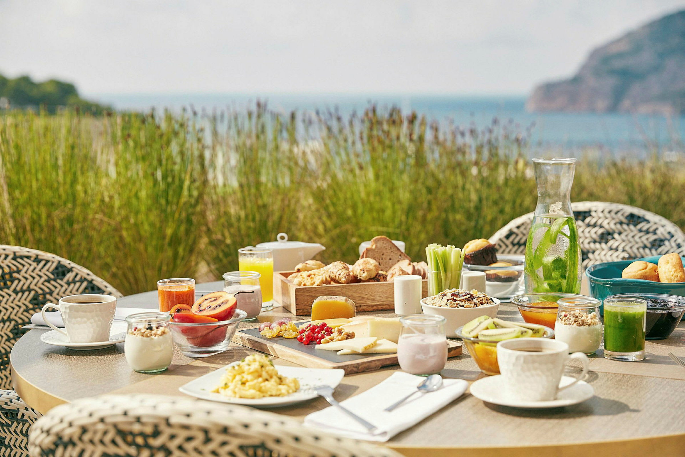 Ett dukat frukostbord utomhus med olika livsmedel och en bakgrund av grönska och hav. (AI-genererad text)