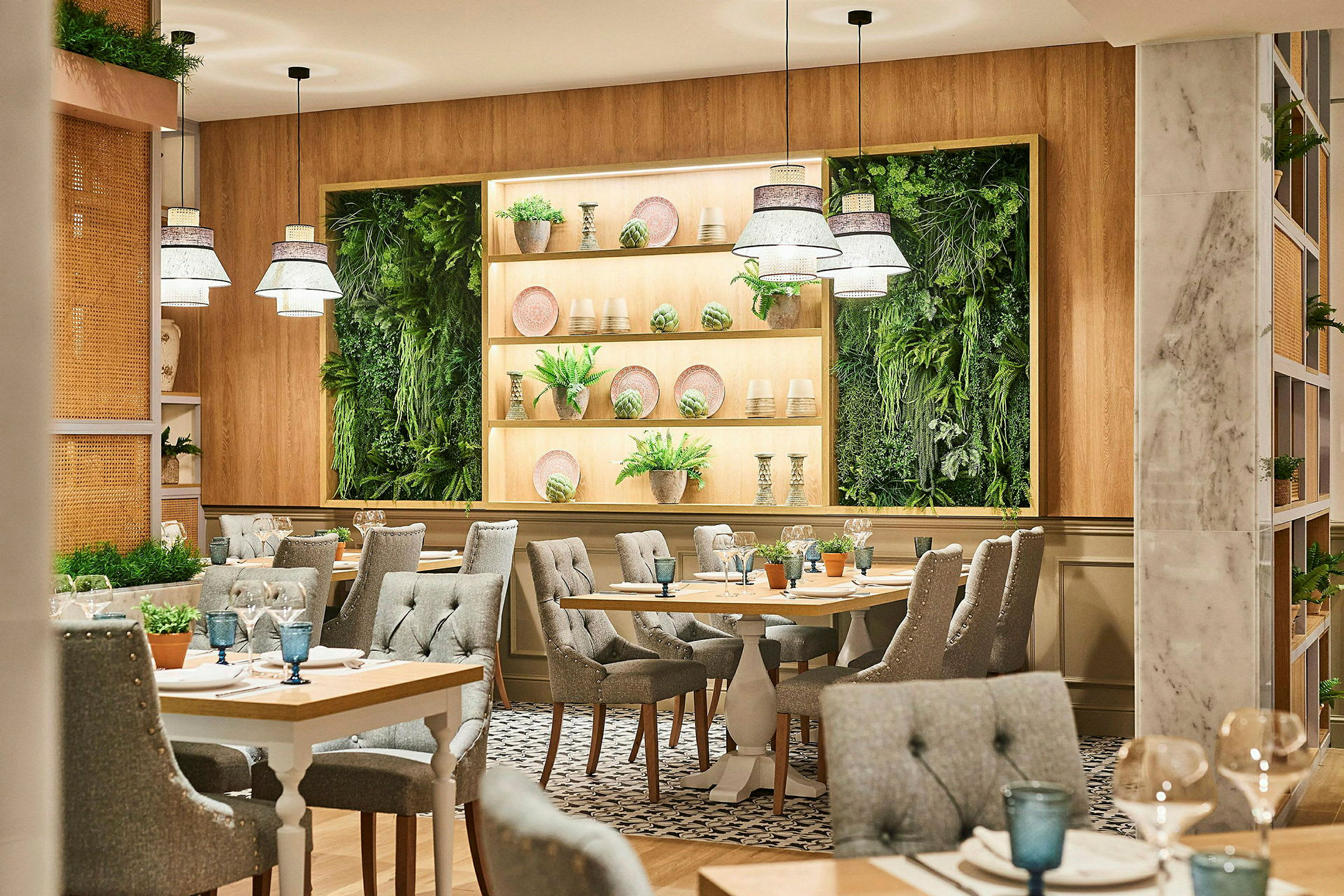 En restaurang med ljus och modern inredning. Bord är omgivna av gråa vadderade stolar, och på väggen finns gröna växtpaneler och hyllor med dekorationer. (AI-genererad text)