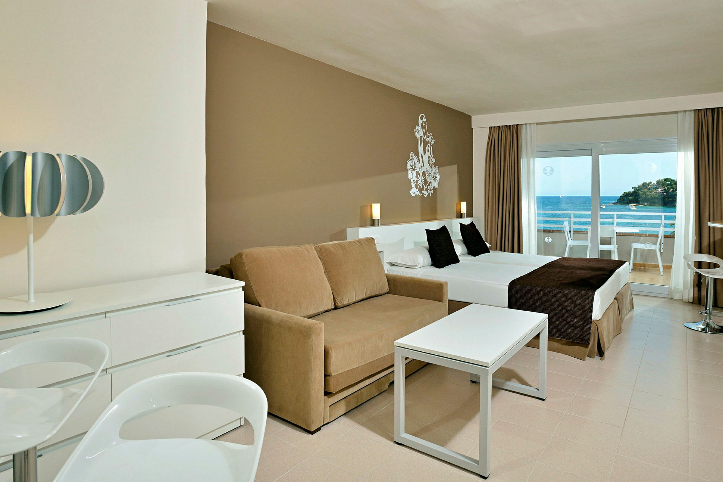 Ett hotellrum med modern inredning, inklusive en soffa, ett bord, en säng och en balkong som vetter mot havet. (AI-genererad text)