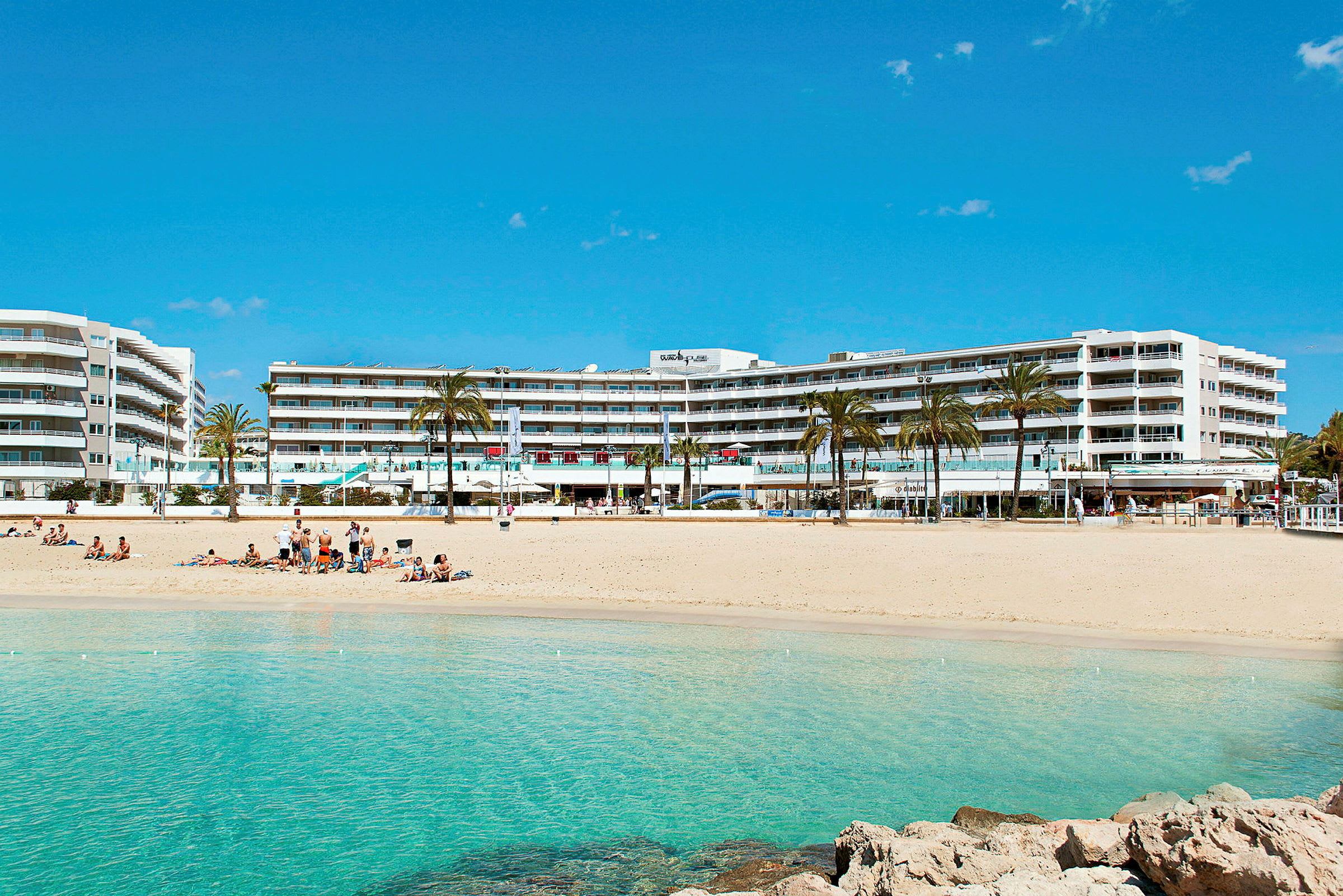 En sandstrand med turkost vatten framför en modern hotellbyggnad omgiven av palmer. (AI-genererad text)