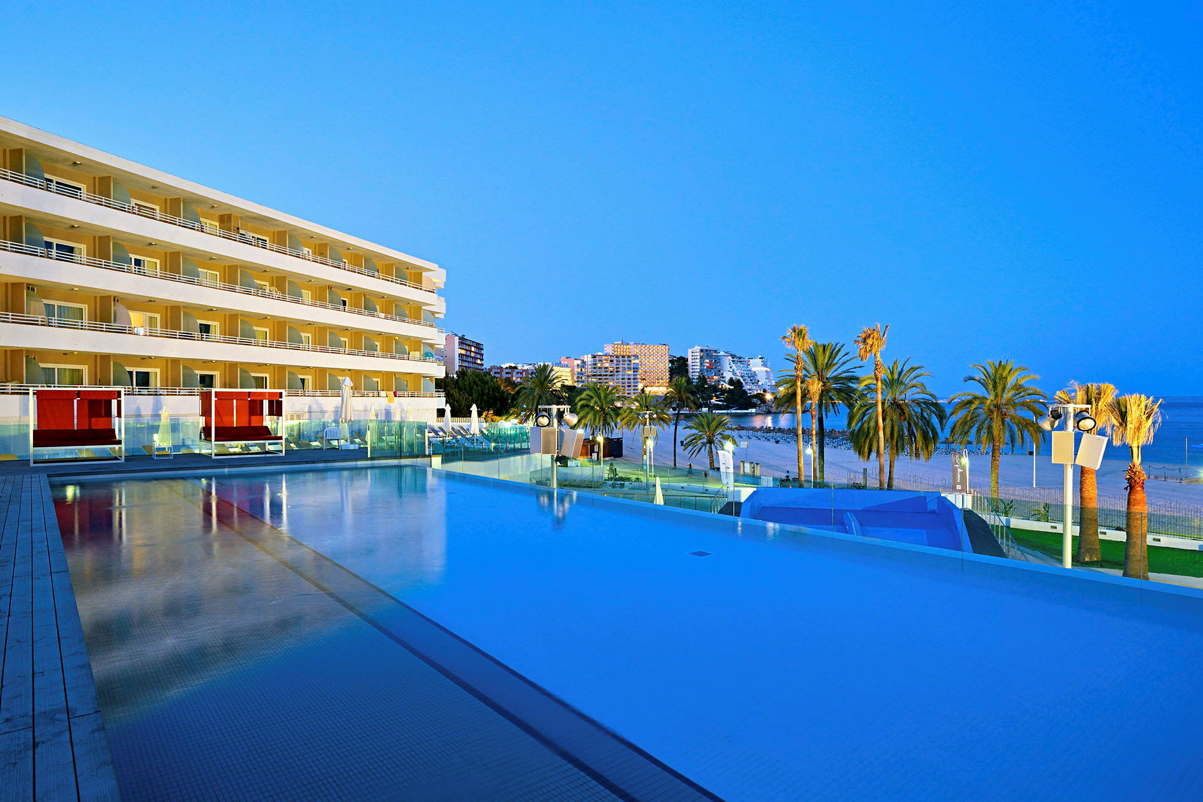 Ett hotell med flera våningar och en pool framför, beläget vid en strand kantad av palmer med havet i bakgrunden. (AI-genererad text)