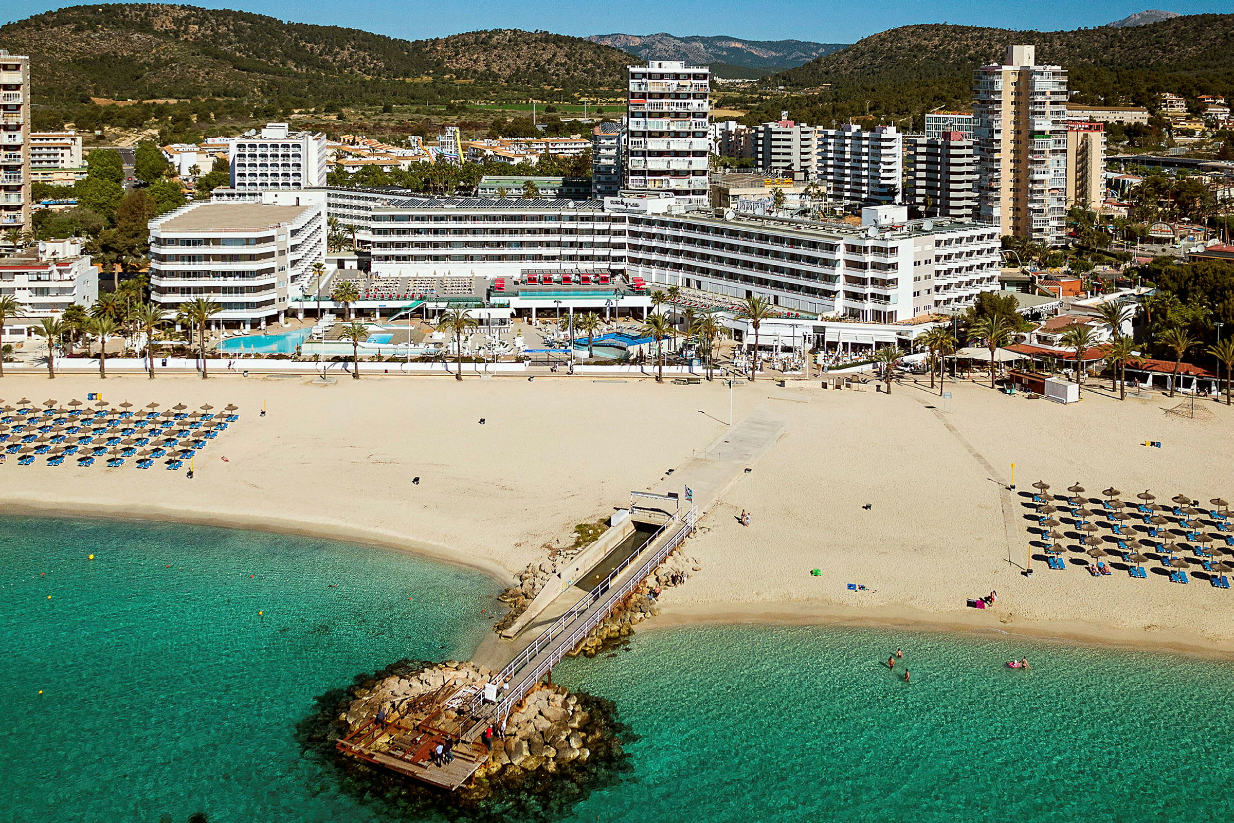 Bild av en sandstrand med solstolar och parasoll, en brygga som leder ut i havet, och hotellbyggnader i bakgrunden. (AI-genererad text)