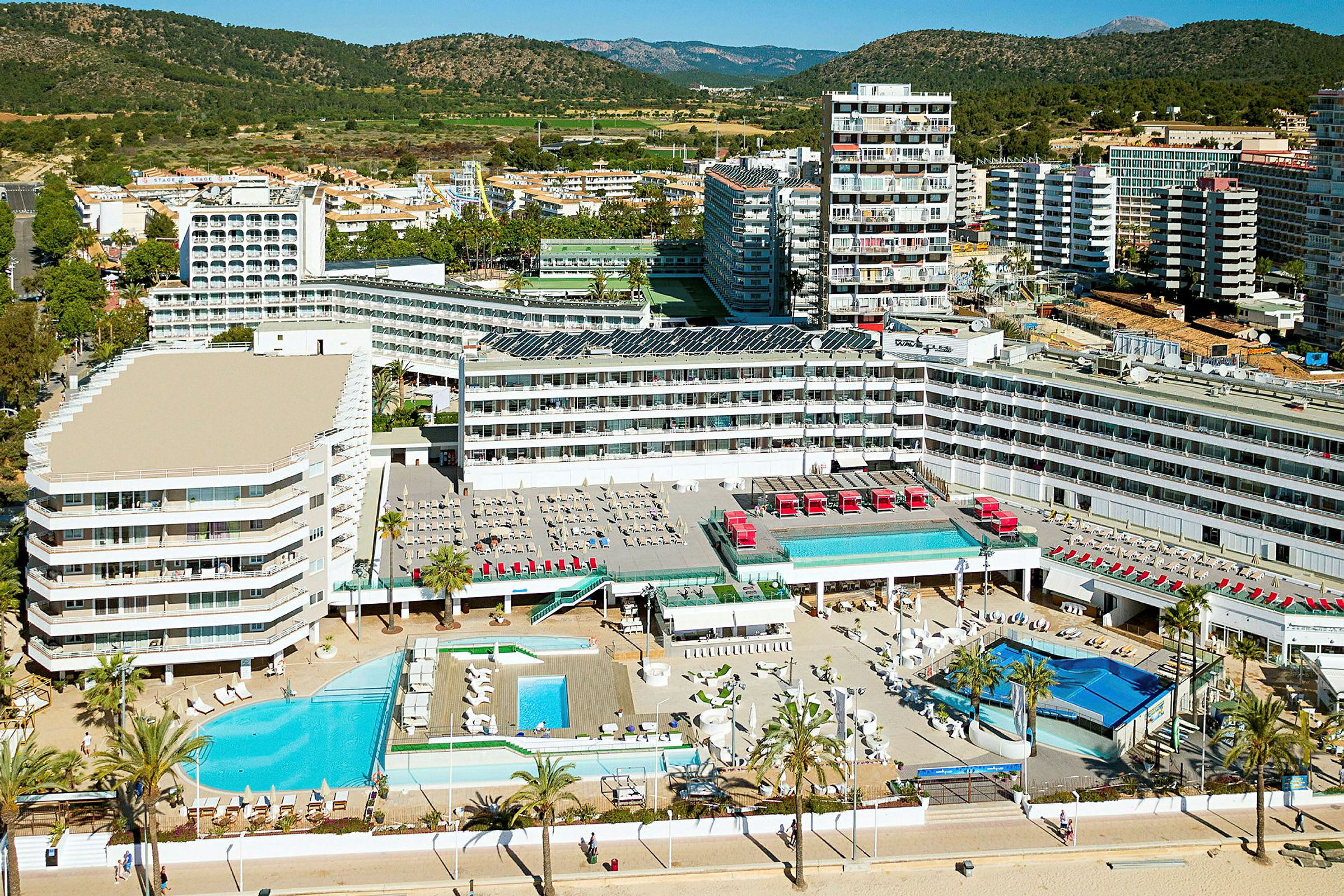 Flygvy över ett hotellkomplex med flera byggnader, pooler och solstolar. Omgivningen inkluderar skog och andra byggnader. (AI-genererad text)