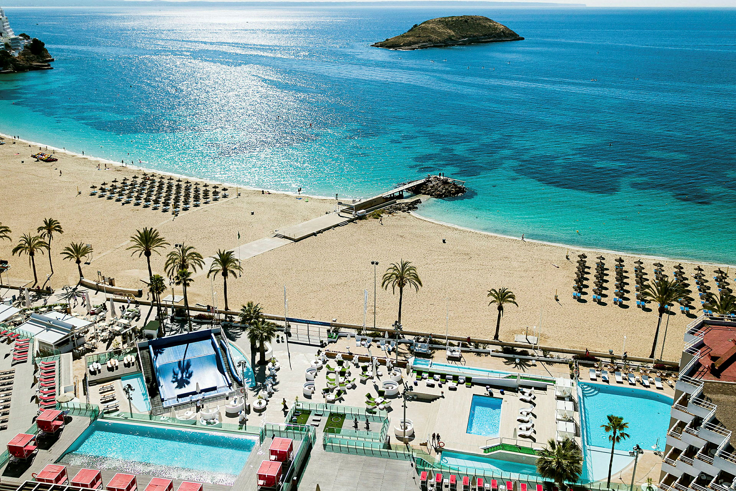 En sandstrand med parasoller och solstolar vid klart blått vatten. I förgrunden en hotellpool och terrass med solstolar. (AI-genererad text)