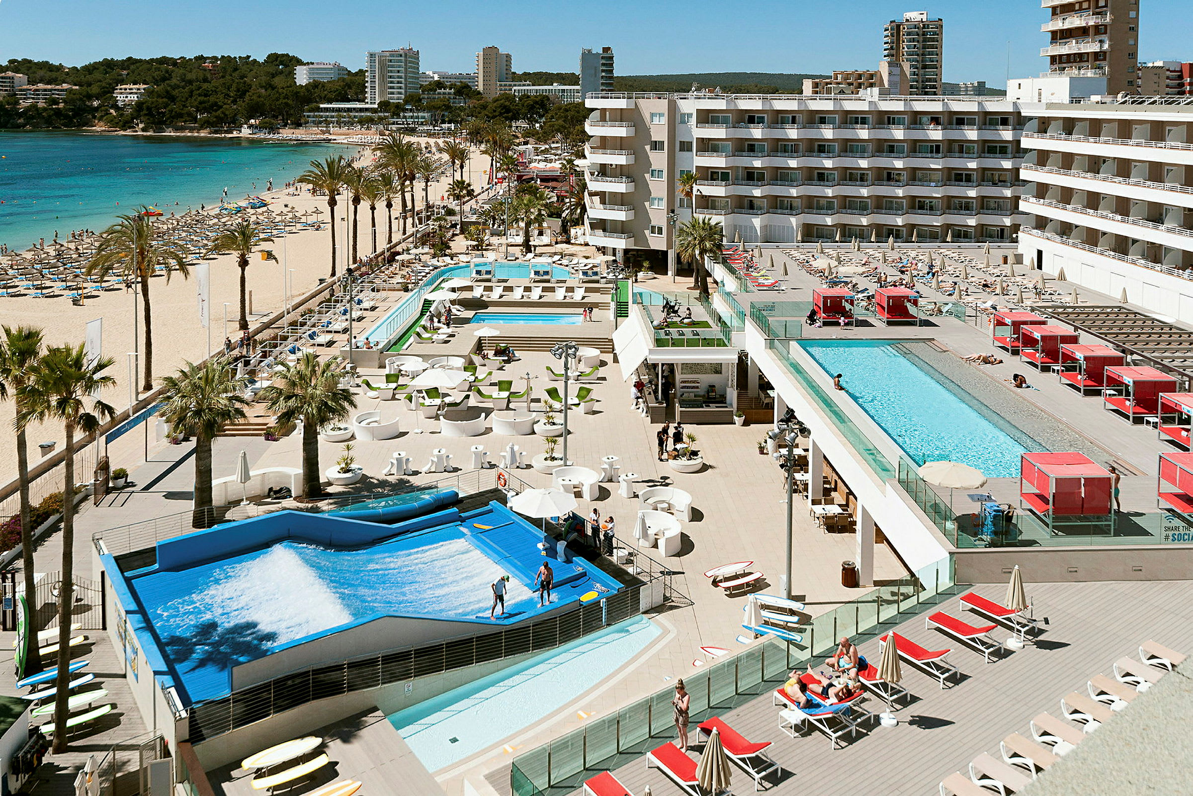 Utsikt över en strand med solsängar och parasoller, omgiven av hotellbyggnader och en stor poolområde. (AI-genererad text)