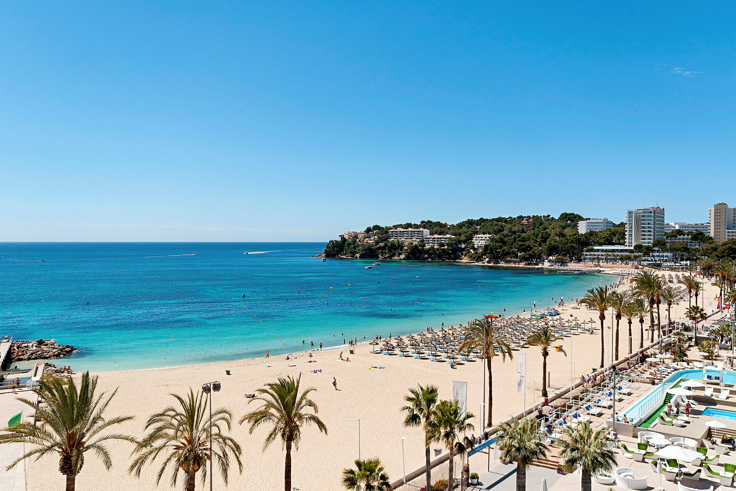 Strand med gyllene sand, palmer, solstolar och parasoller, samt klart blått hav, med hotellbyggnader i bakgrunden. (AI-genererad text)