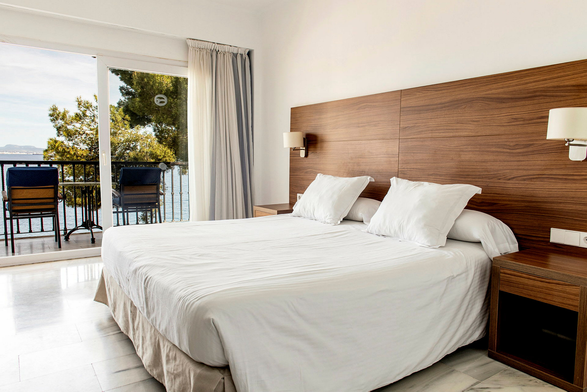 Ett hotellrum med en dubbelsäng, träpanel på väggen och utsikt mot en balkong med hav i bakgrunden. (AI-genererad text)