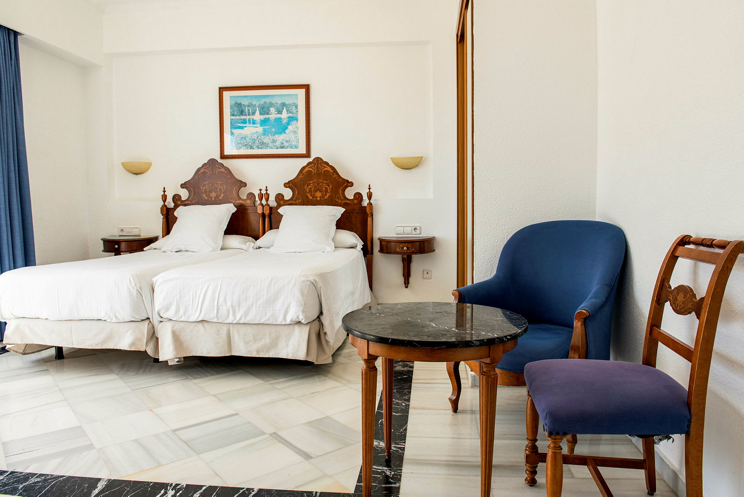 Ett hotellrum med två enkelsängar, en blå fåtölj och en trästol i klassisk stil. (AI-genererad text)