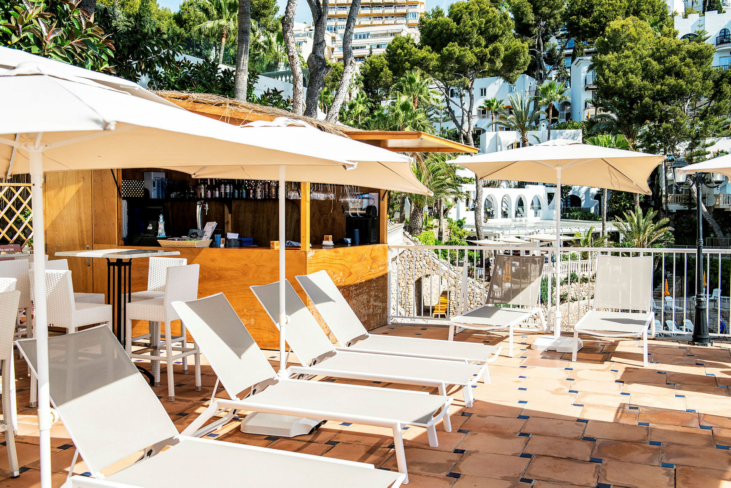 Solsängar och parasoller på en terrass vid en bar, med utsikt över träd och byggnader. (AI-genererad text)