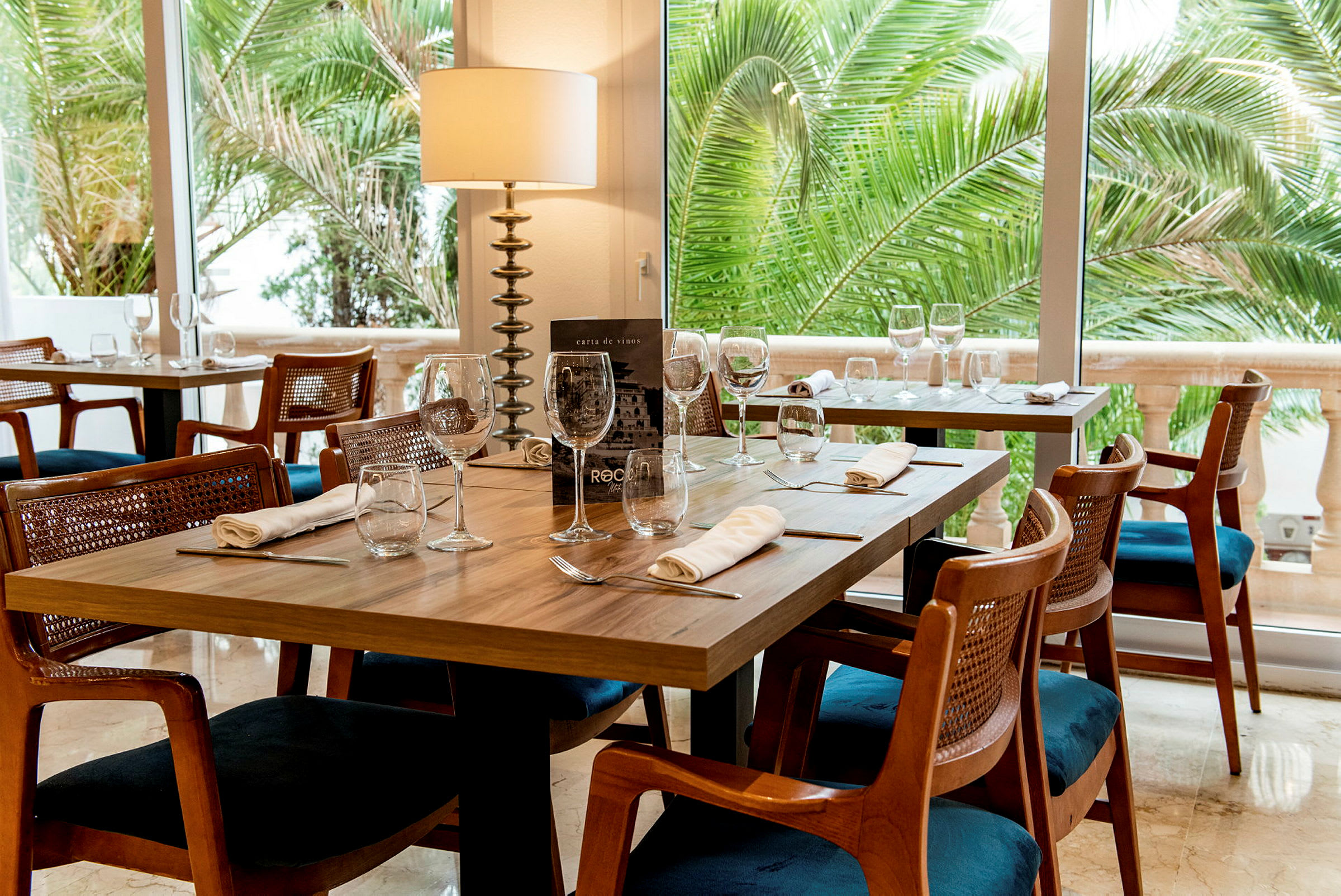Ett restaurangbord med dukning och glas, omgiven av trästolar och gröna palmer synliga genom fönstret. (AI-genererad text)