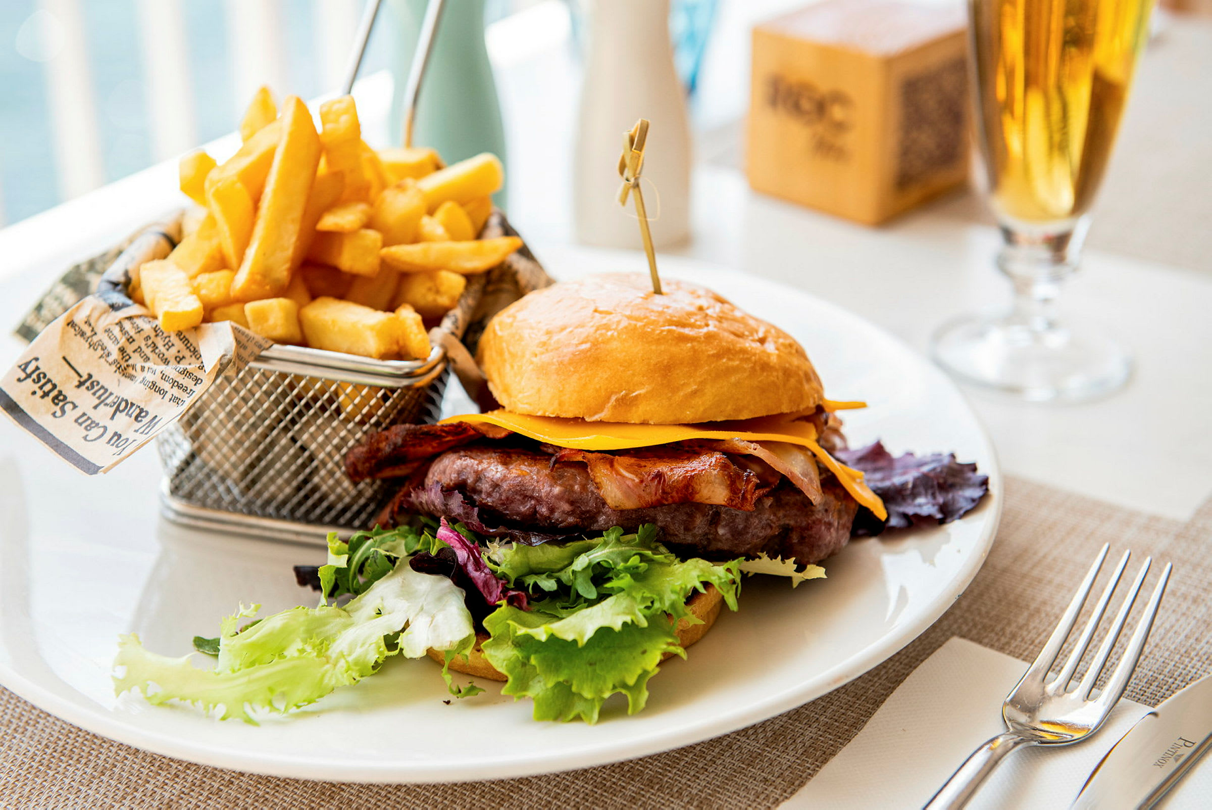 En tallrik med en hamburgare med ost, bacon och sallad, samt en korg med pommes frites. (AI-genererad text)