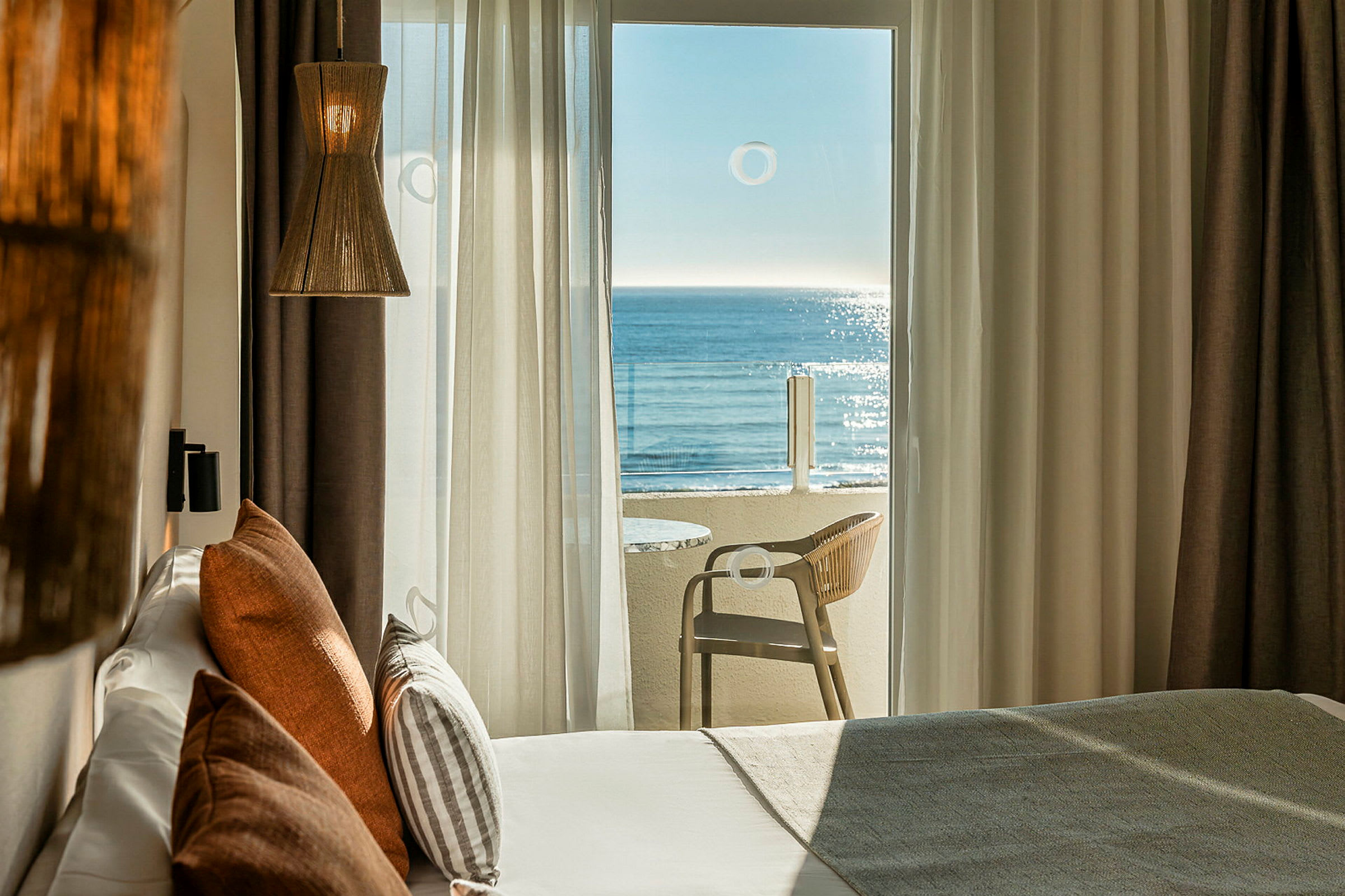 Ett hotellrum med en stor balkongdörr som leder till en balkong med utsikt över havet. Sängen är utrustad med dekorativa kuddar och en lampa hänger från taket. (AI-genererad text)