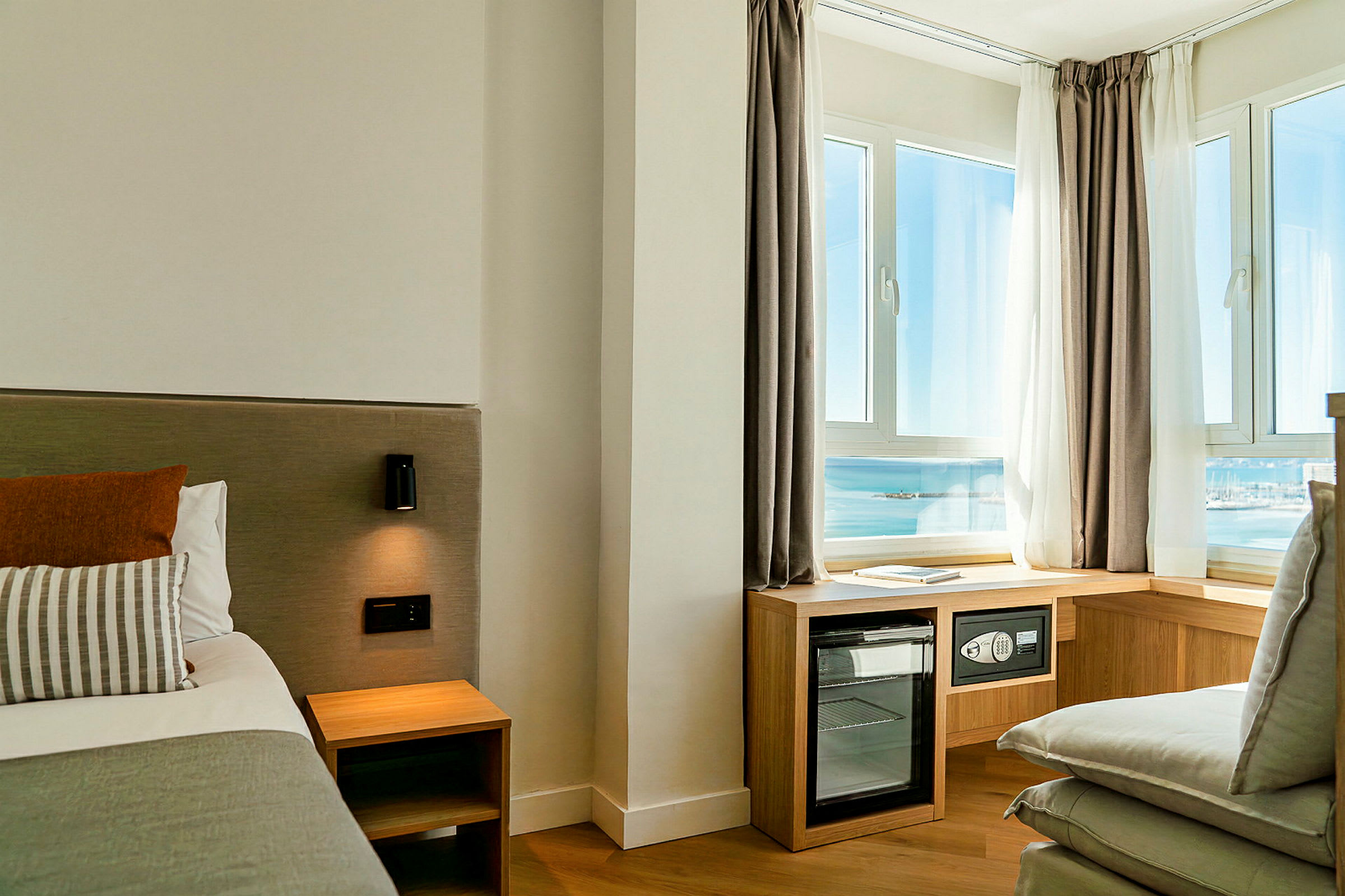 Hotellrum med del av en säng, nattbord, vägglampa och stora fönster med utsikt mot havet samt möbler som ett minibarkylskåp. (AI-genererad text)