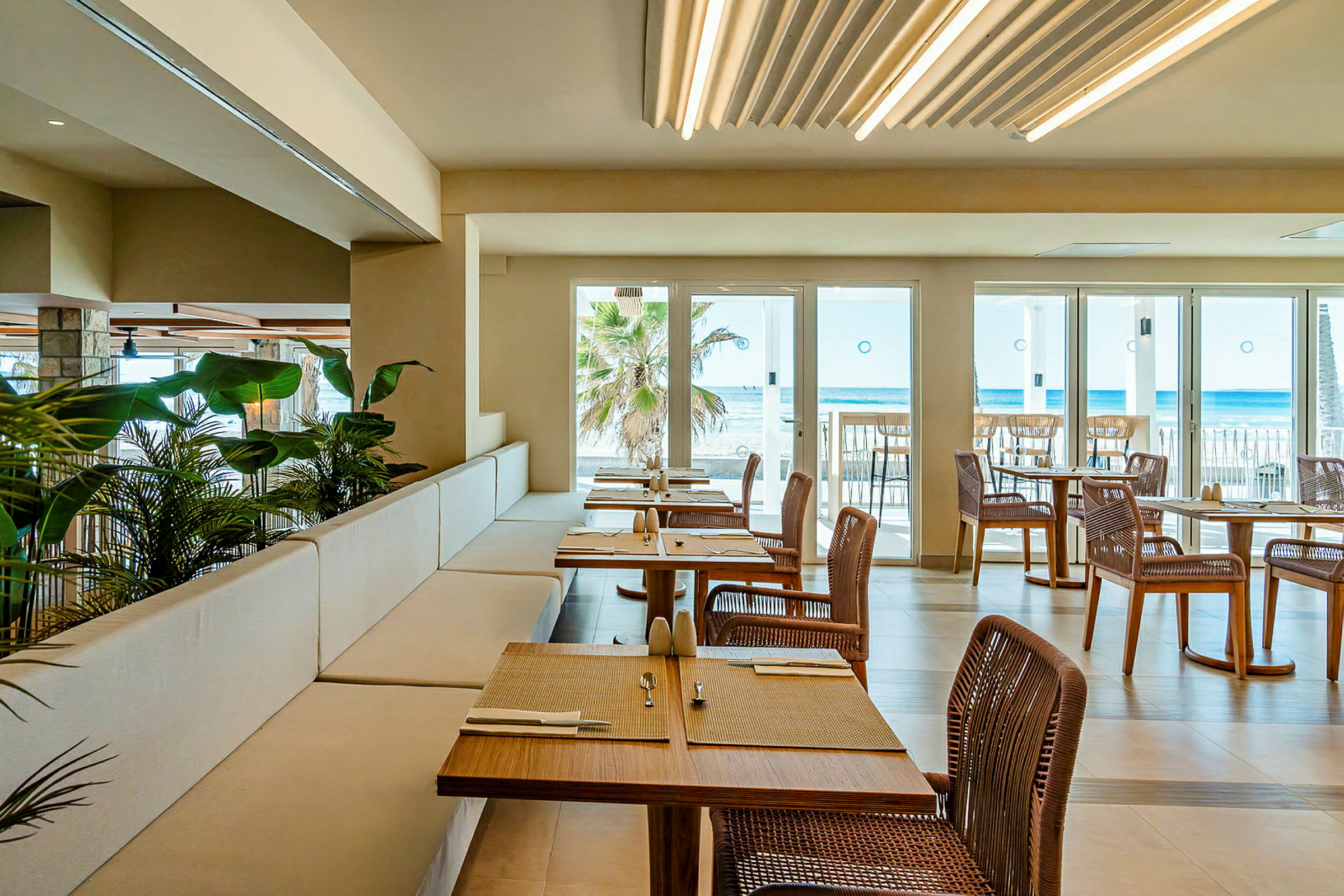 En restaurang med utsikt över havet. Borden är dukade och stolar i rotting är arrangerade runt dem. Stora glasfönster och dörrar släpper in naturligt ljus. (AI-genererad text)