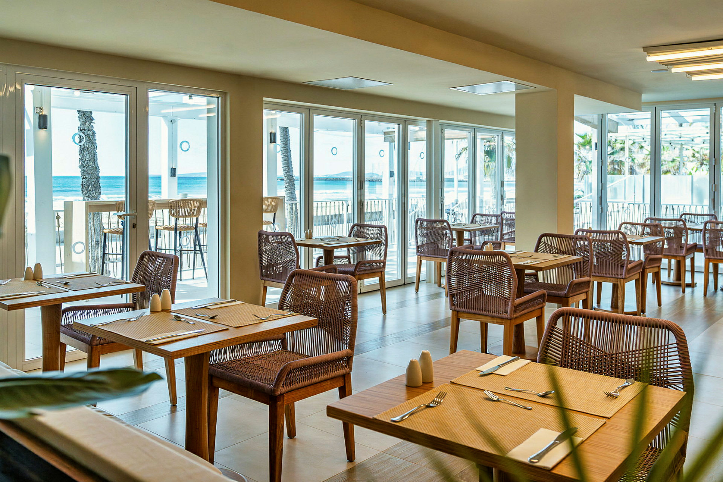 En restaurang med träbord och flätade stolar, med havsutsikt genom glasfönster. (AI-genererad text)