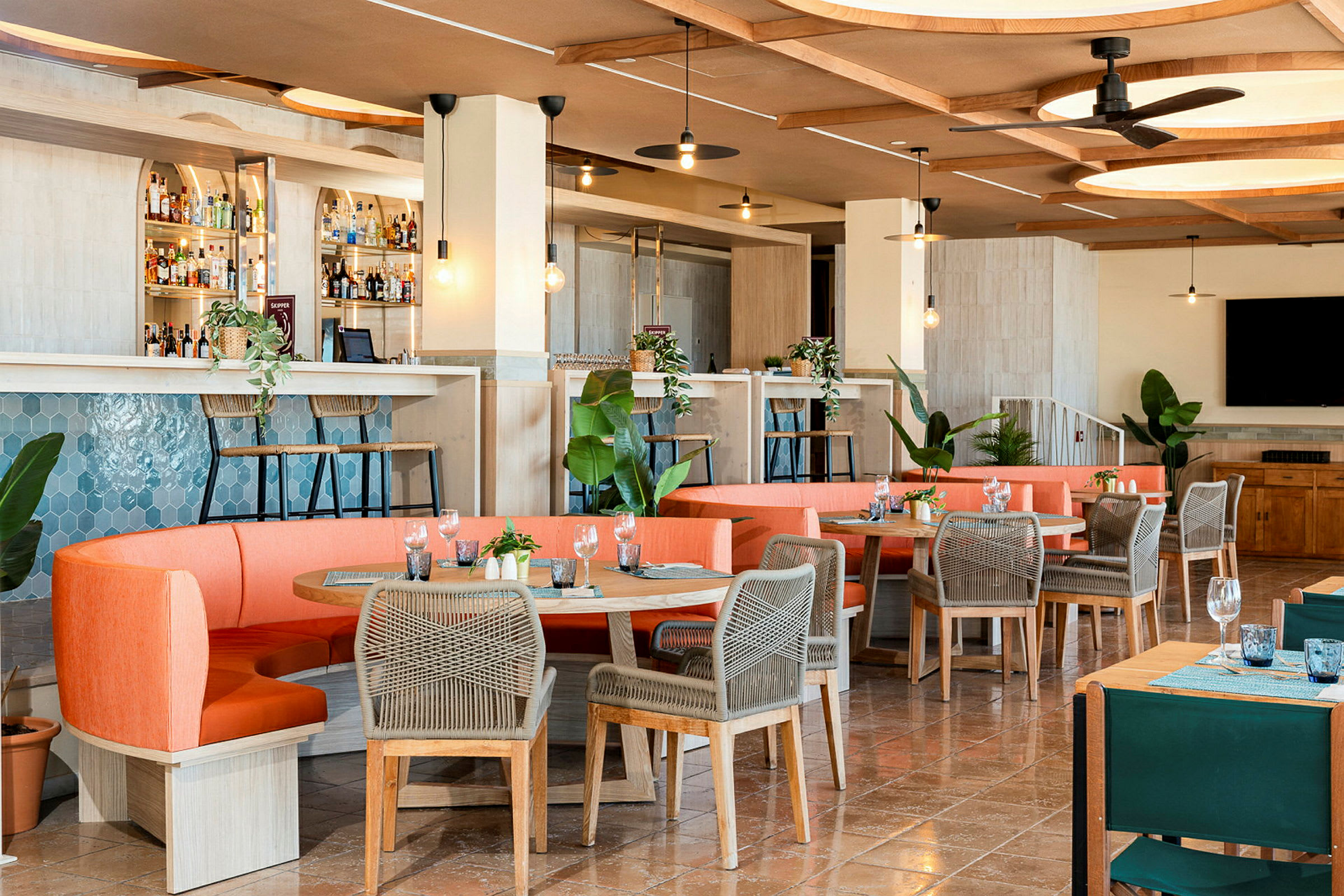 En modern restaurang med välkomnande inredning, orangea soffor och bord med grå stolar, samt en bardisk och gröna växter. (AI-genererad text)