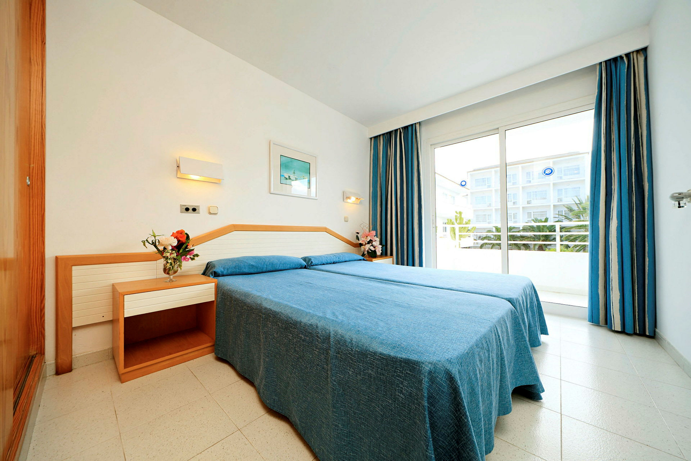 Ett ljust hotellrum med en dubbelsäng som har blå sängkläder, två nattduksbord med dekorationer och en balkong med utsikt. (AI-genererad text)