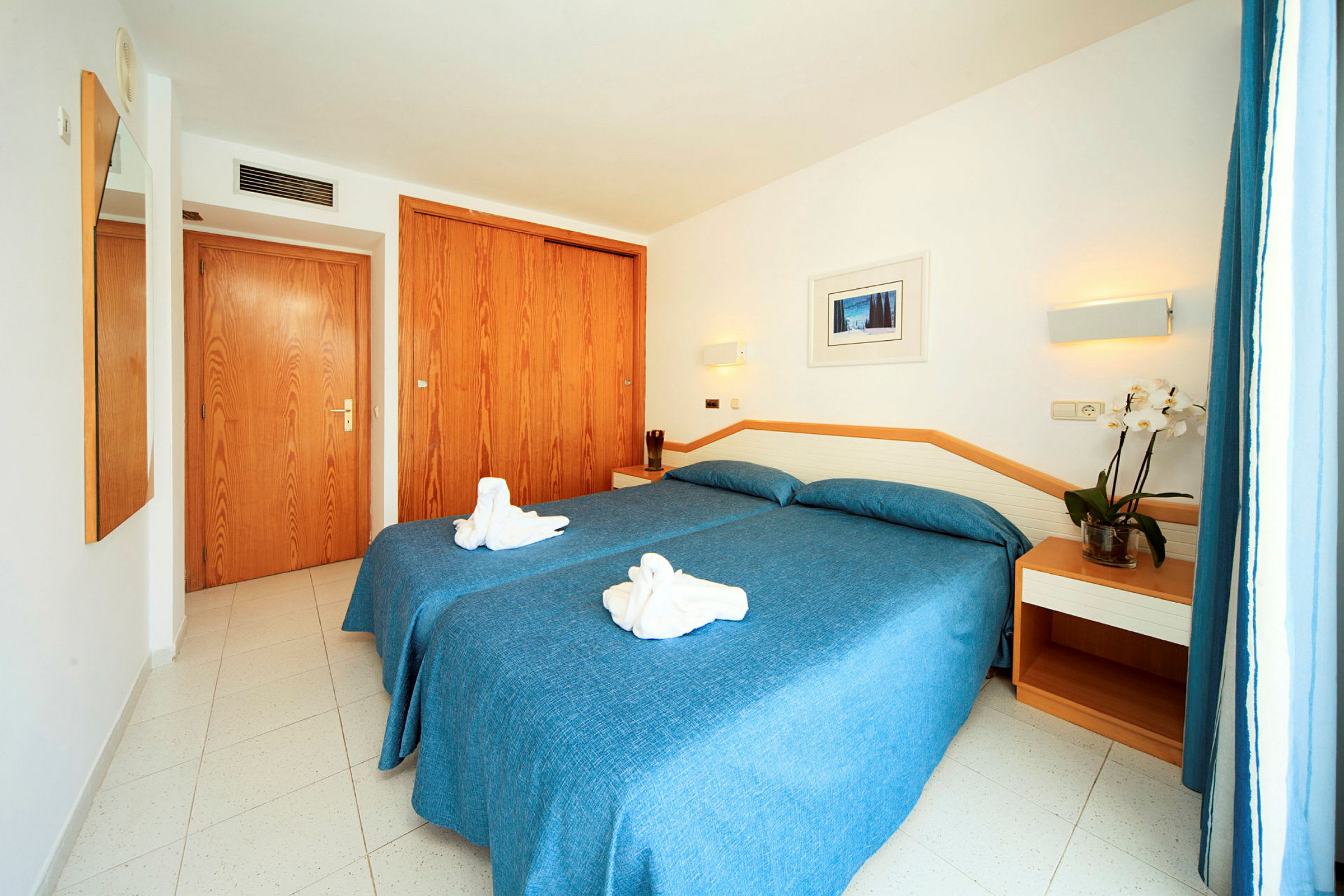 Hotellrum med två sängar bäddade med blåa sängkläder, en växt på nattduksbordet och handdukar vikta som dekorativa figurer. (AI-genererad text)
