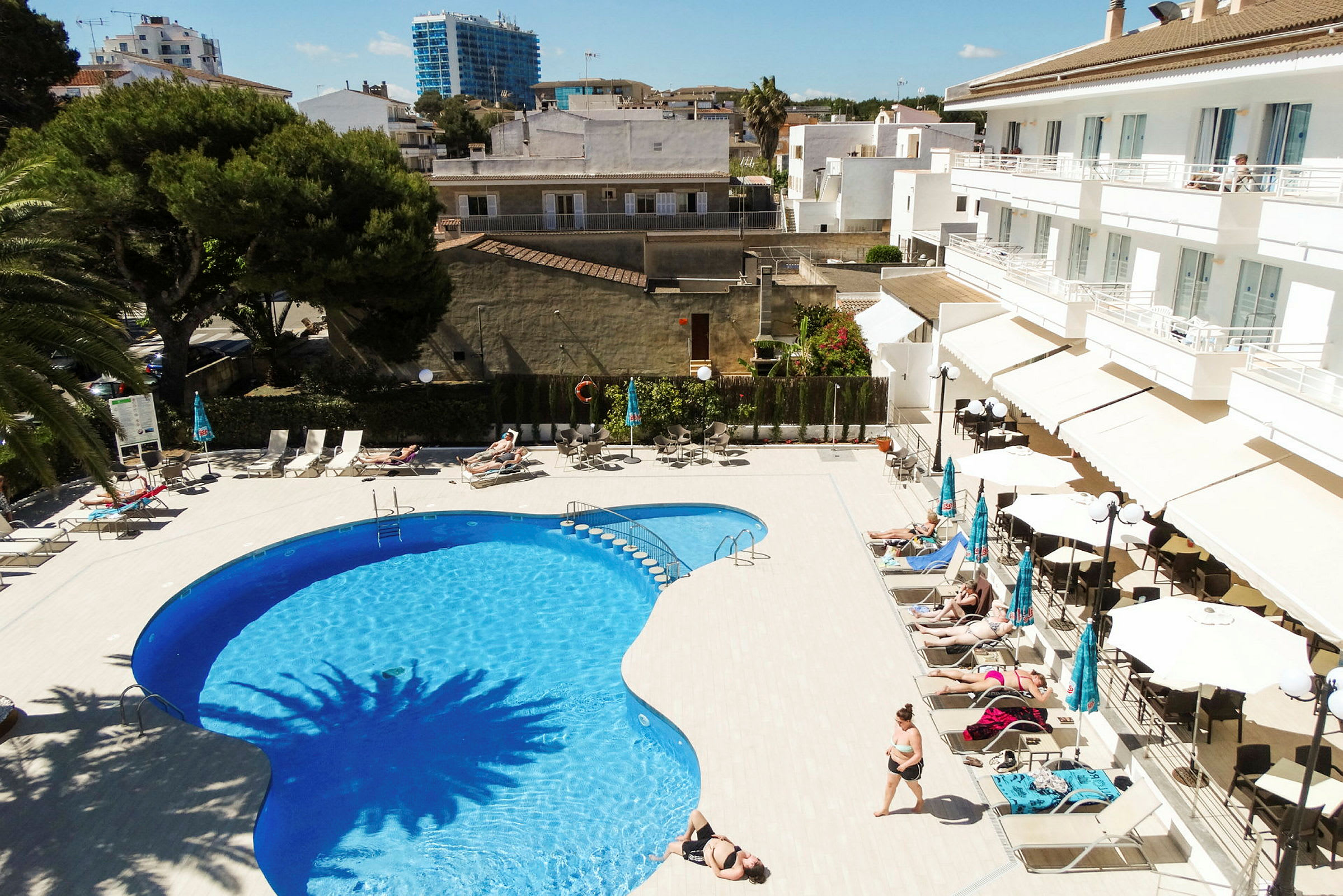 Utomhuspool med solstolar och parasoller vid sidan, flankerad av ett hotell och omgivande byggnader. (AI-genererad text)