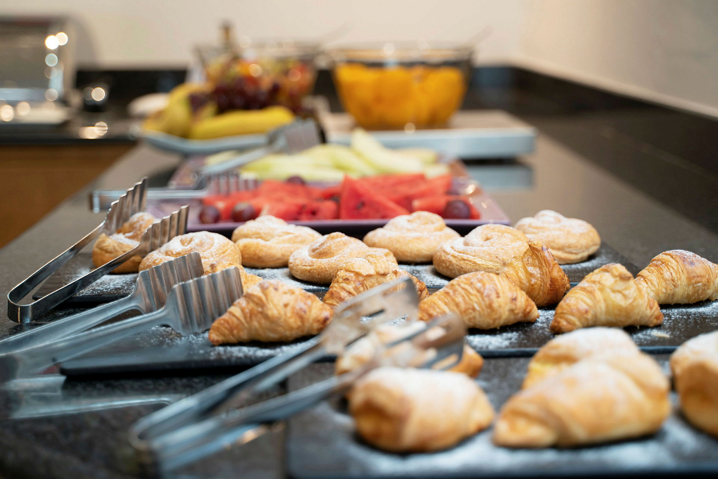 Ett buffébord med croissanter och andra bakverk framställda bredvid tänger för servering. I bakgrunden syns färsk frukt. (AI-genererad text)