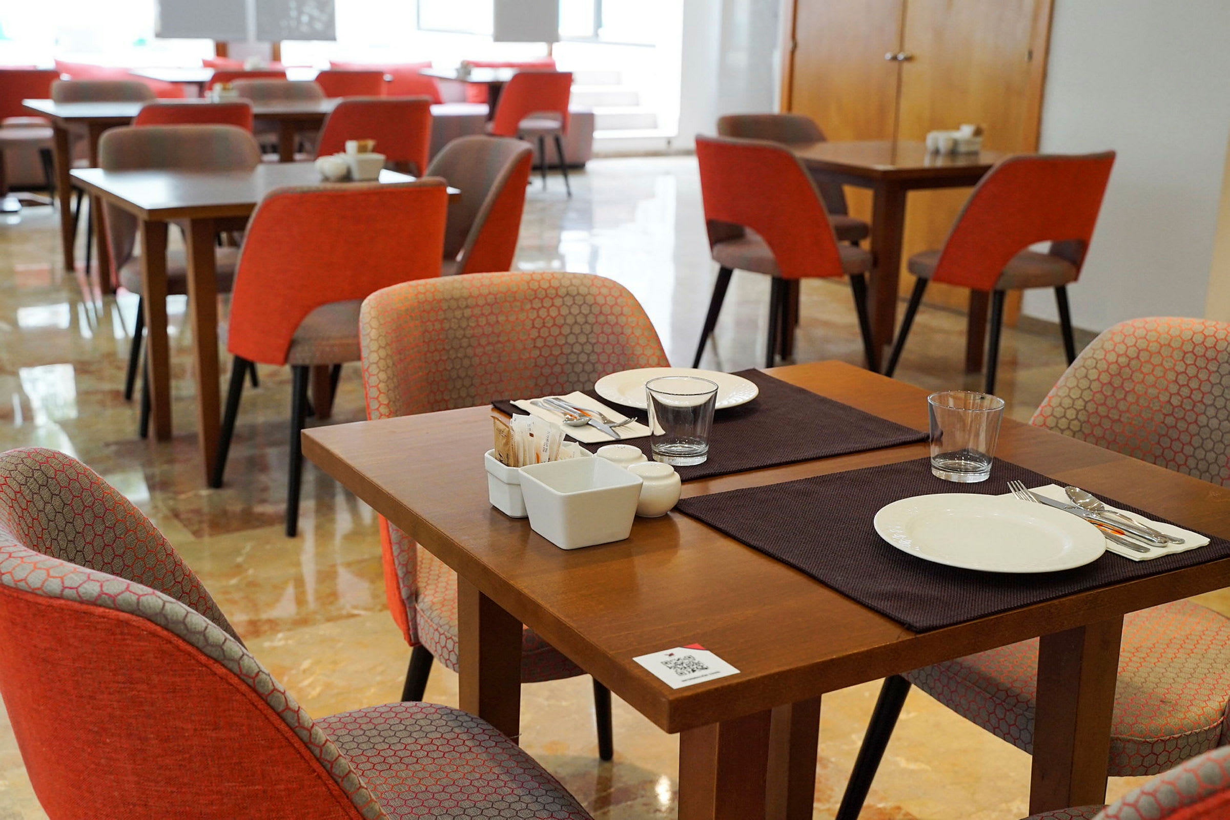 Ett inomhuskafé eller restaurang med träbord och orangea stolar, dukade för matservering. (AI-genererad text)