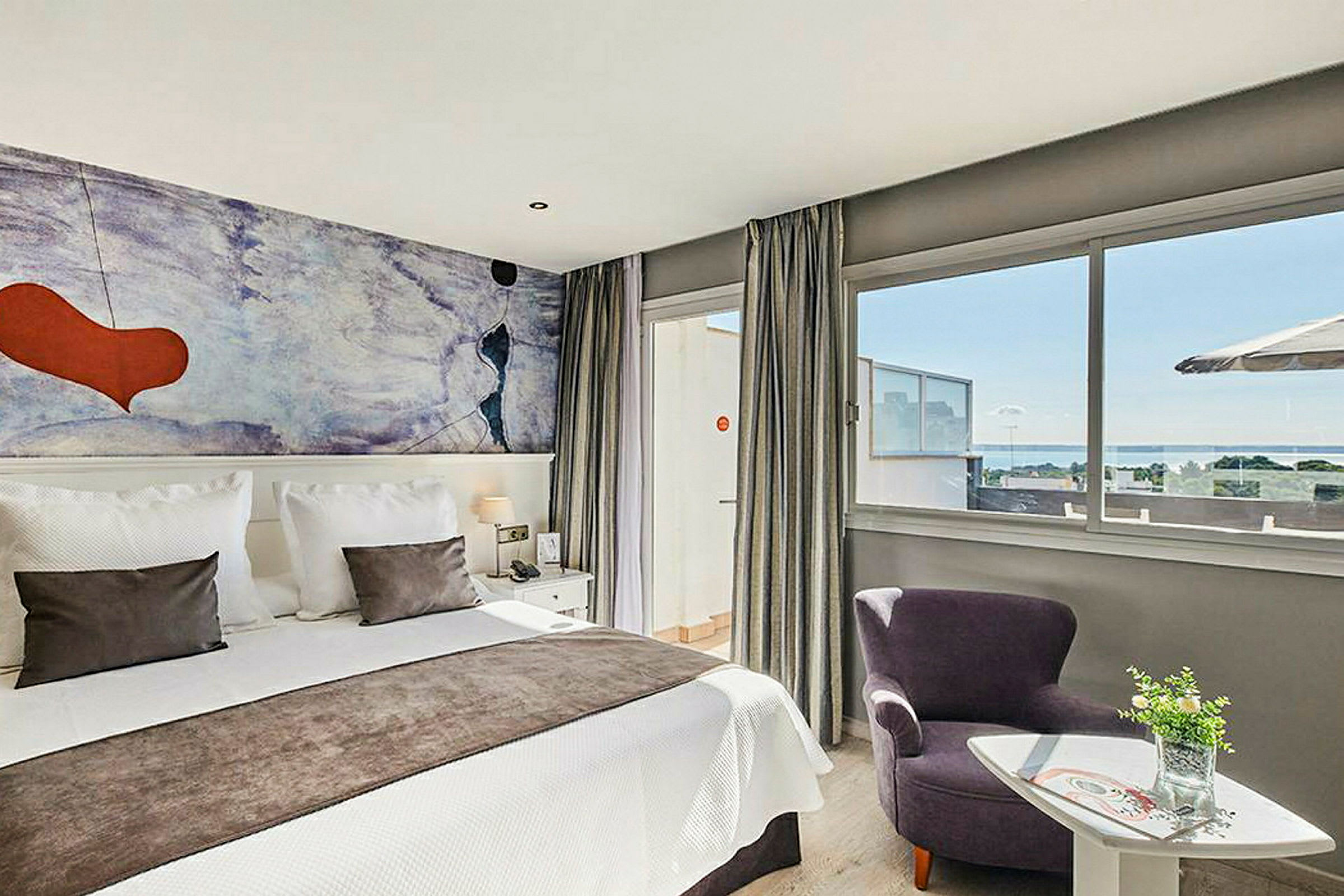 Ett modernt hotellrum med en dubbelsäng, färgglad väggmålning och stora fönster med utsikt över havet. (AI-genererad text)