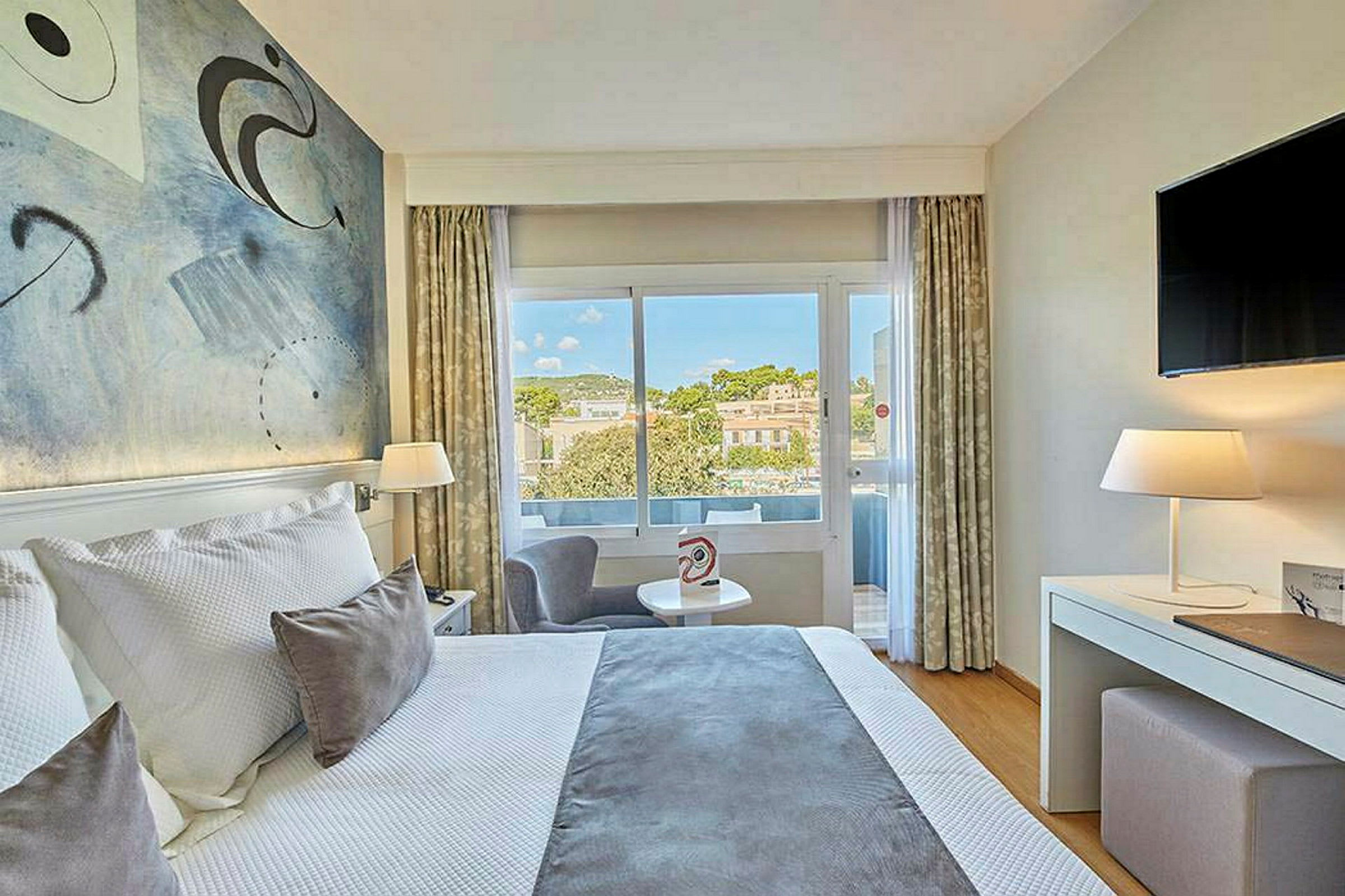 Ett hotellrum med en stor säng, dekorativ konst på väggen, och ett fönster med utsikt över grönska och byggnader. (AI-genererad text)